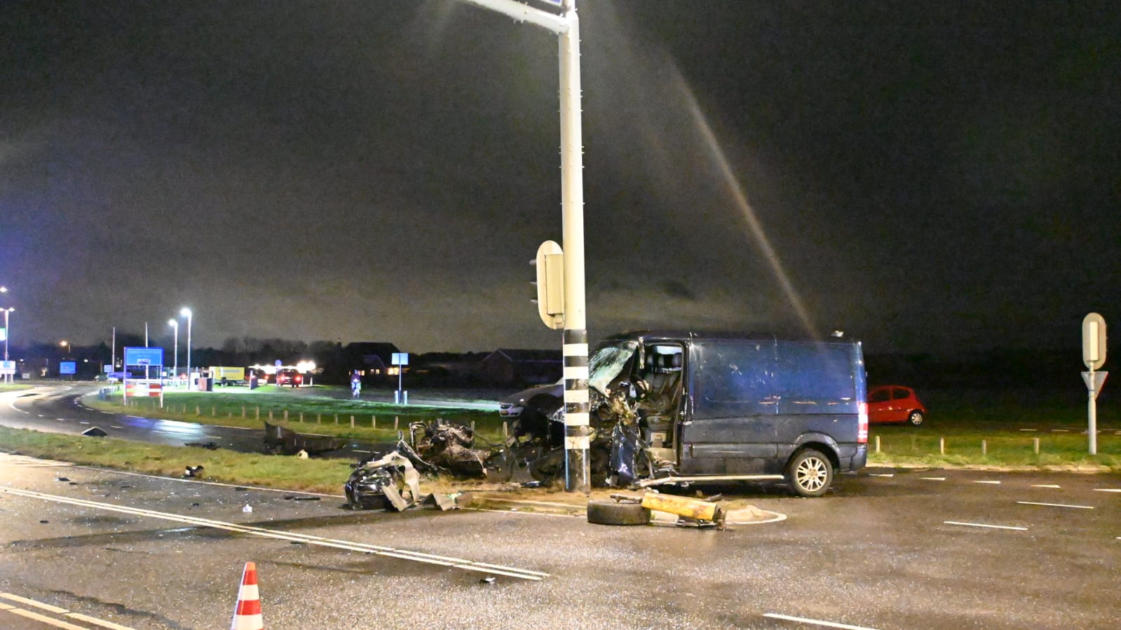 Nachtelijk ongeval waarbij een blauwe bestelbus tegen een lantaarnpaal is gebotst op een kruising, omringd door brokstukken en een verkeerskegel.