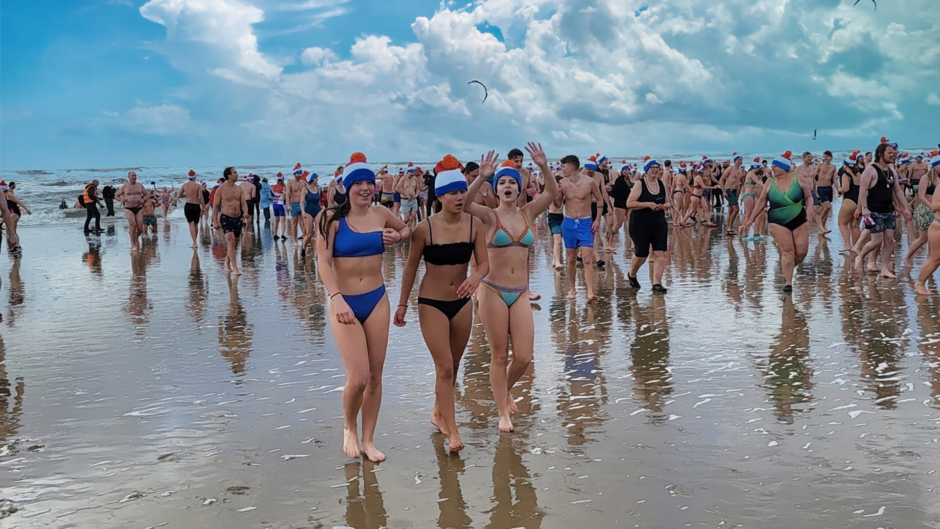 Mensen dragen badkleding en kerstmutsen, wandelen op een druk strand met bewolkte lucht.