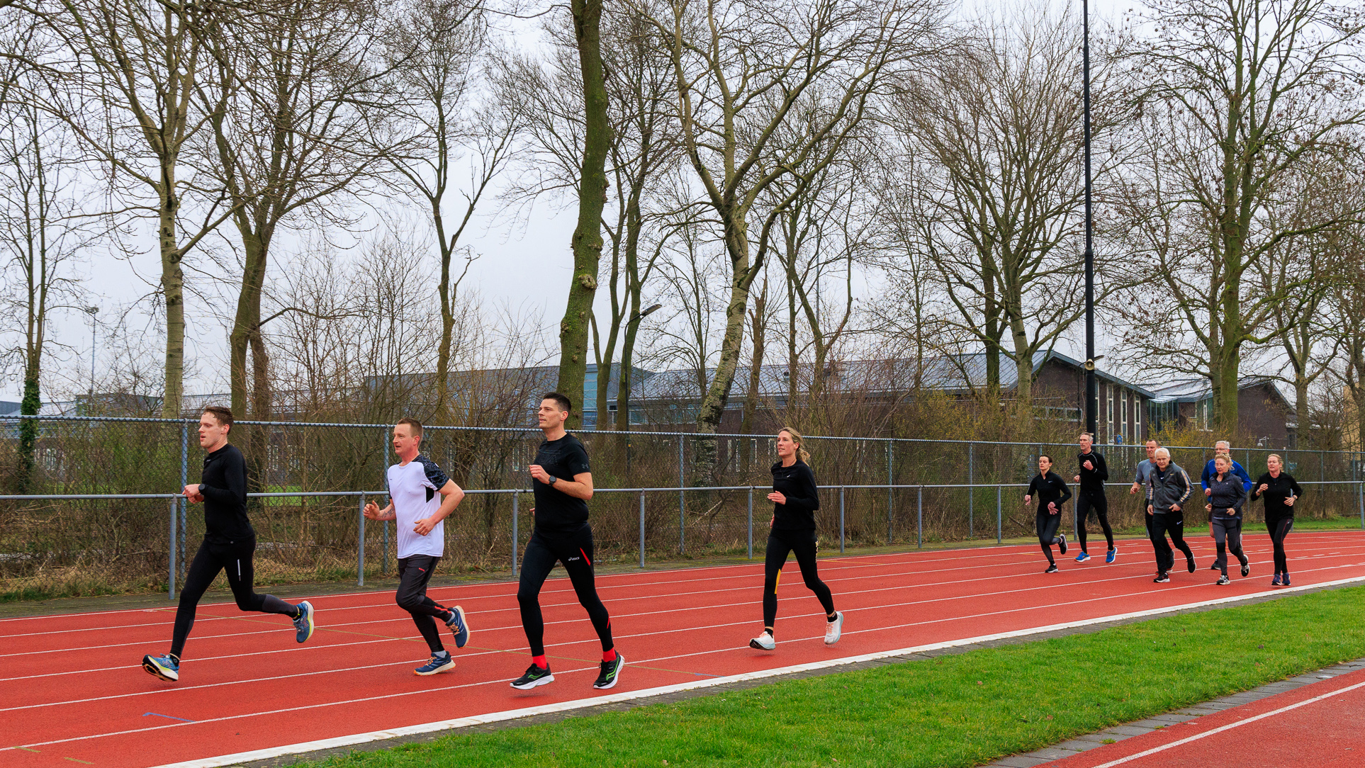 Een groep mensen rent op een atletiekbaan, omgeven door kale bomen op een grijze dag.