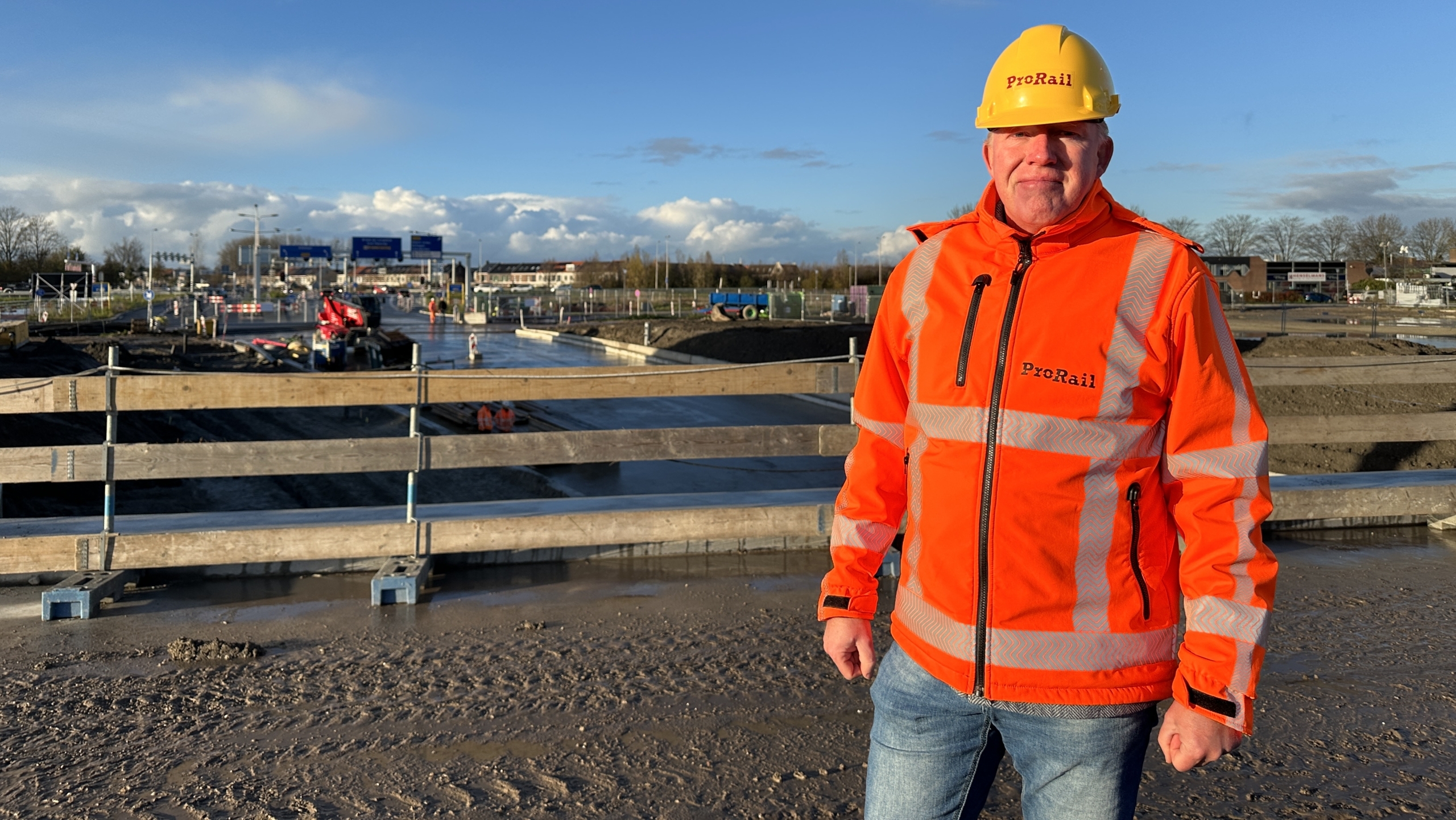Een persoon met een oranje ProRail-jas en gele helm staat op een bouwplaats bij een spoorwegproject, met bouwmachines en een heldere lucht op de achtergrond.