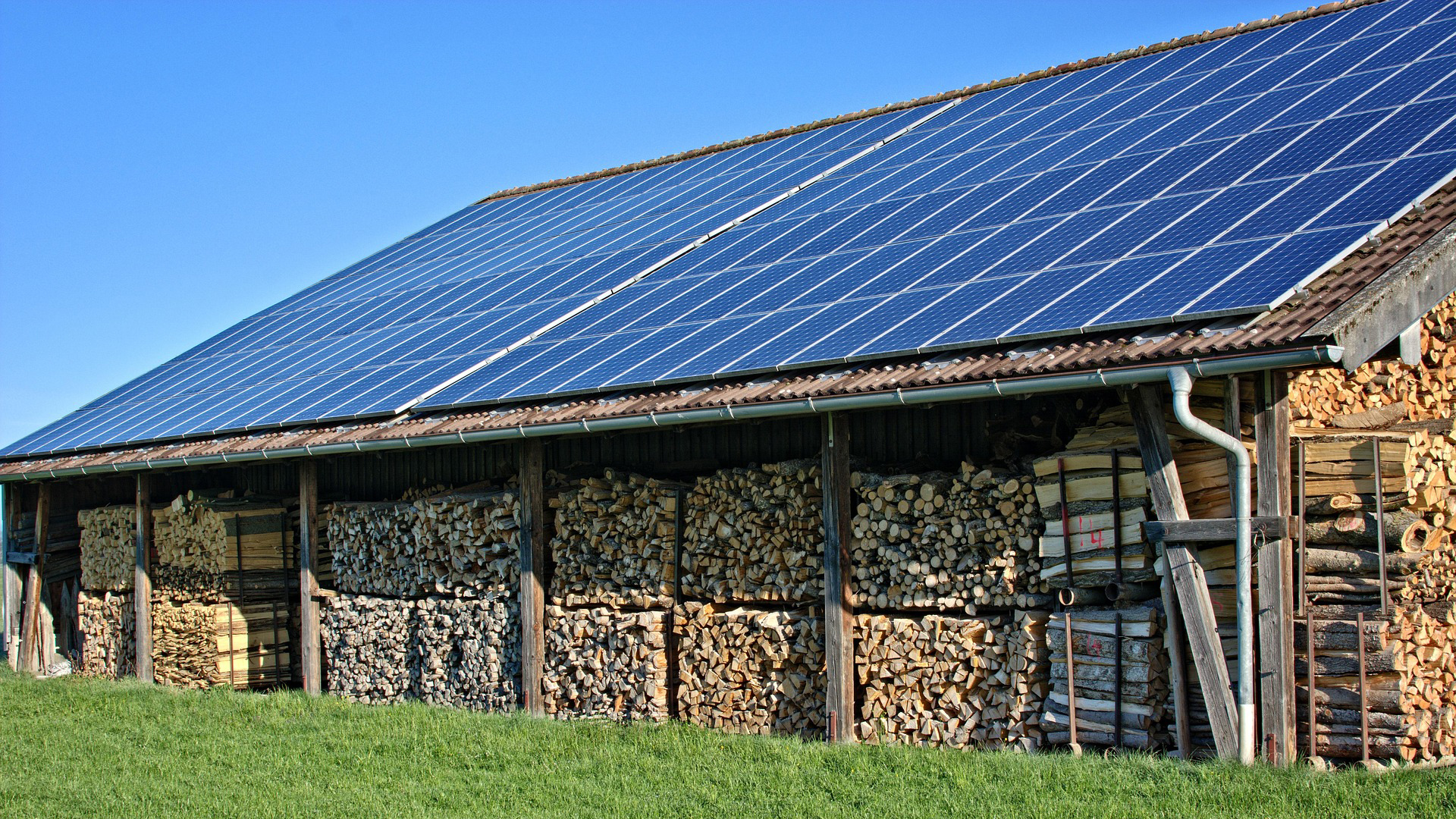 Opslagplaats voor hout met zonnepanelen op het dak, gelegen op een grasveld onder een blauwe hemel.