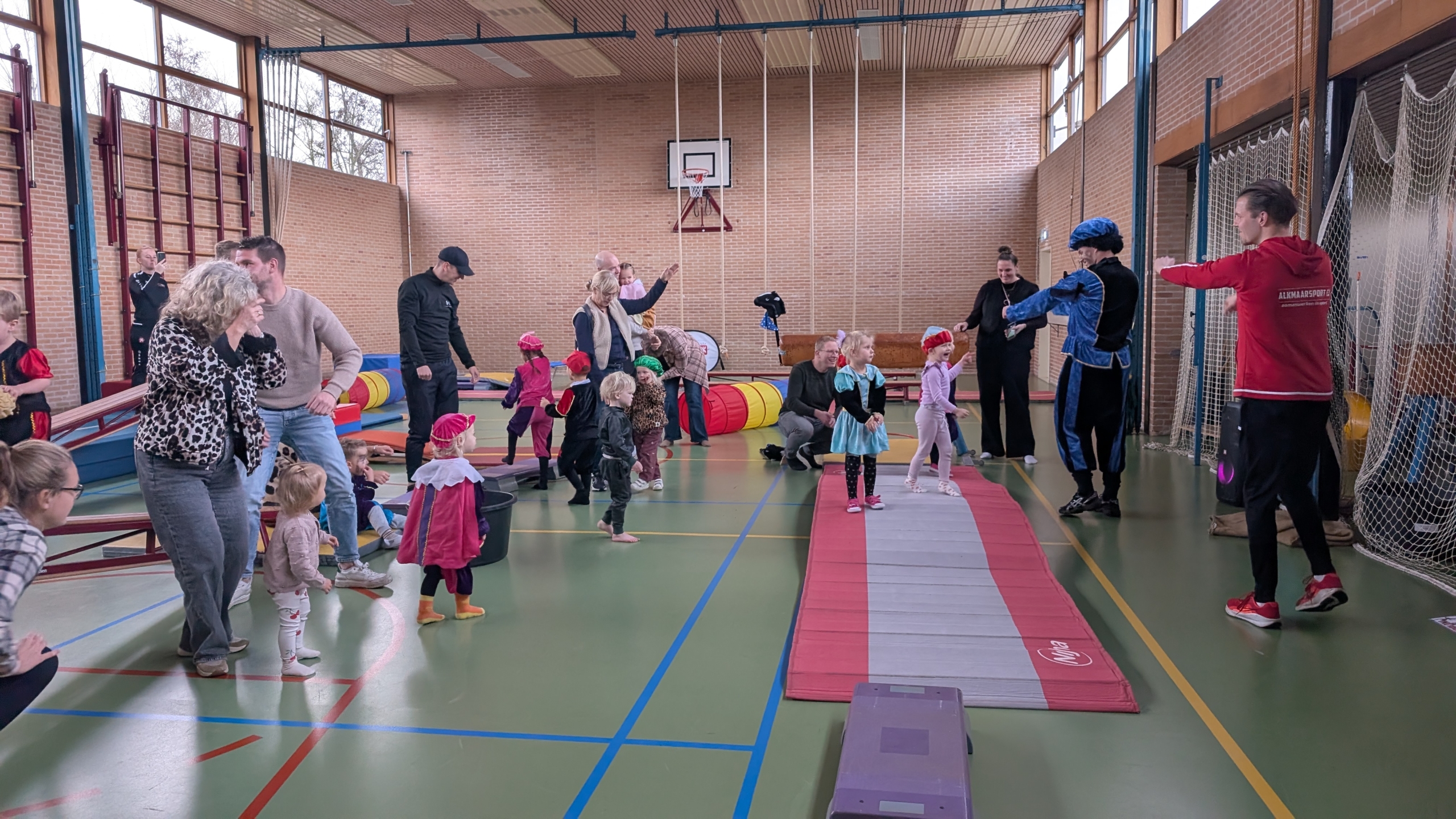 Kinderen en volwassenen doen mee aan een activiteit in een gymzaal, waarbij sommige mensen verkleed zijn, met gymmateriaal zoals matten en tunnels in de ruimte.