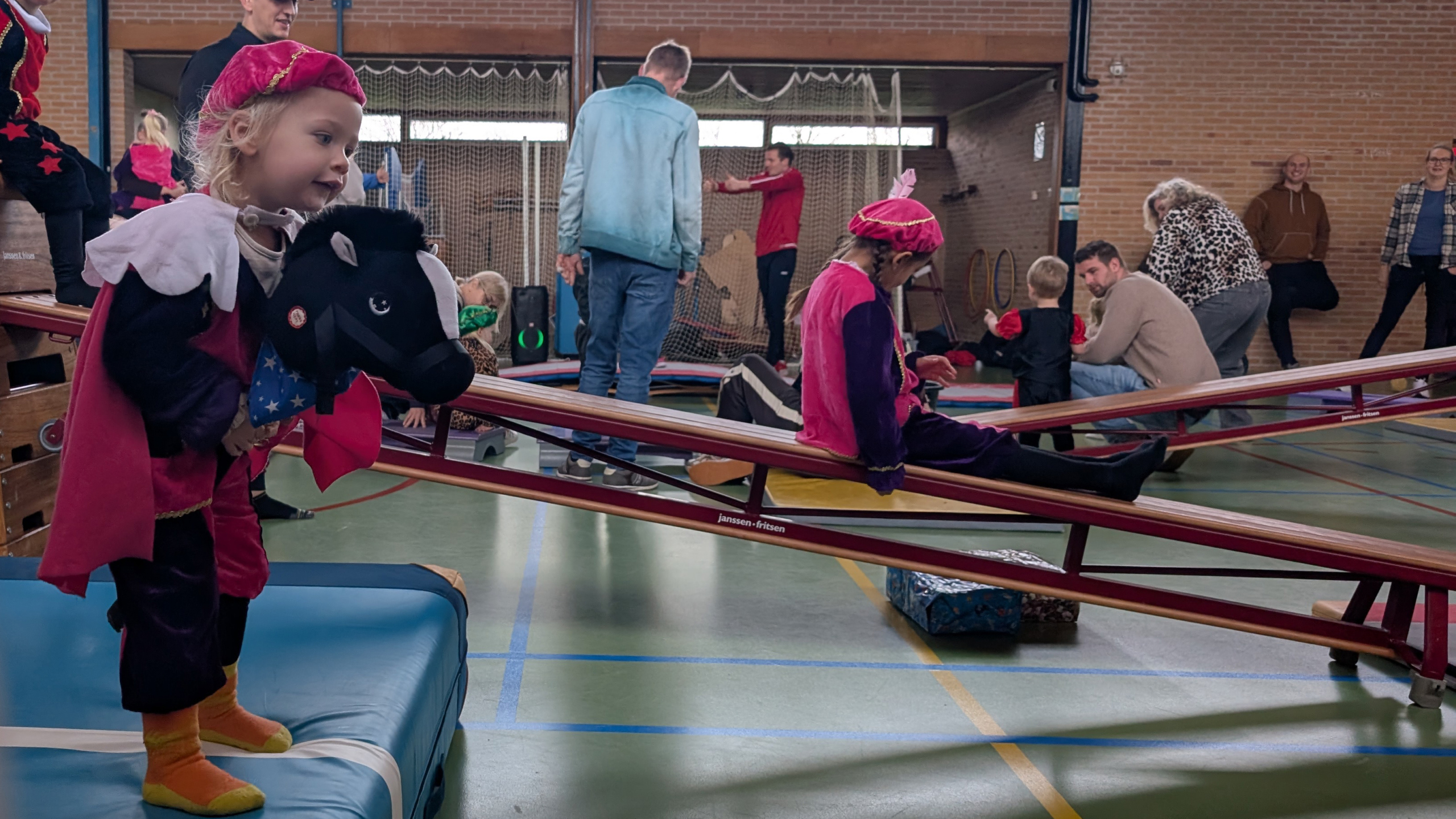 Kinderen in Piet-kostuums spelen in een gymzaal, met een kind op een schommelpaard en anderen op een wip.