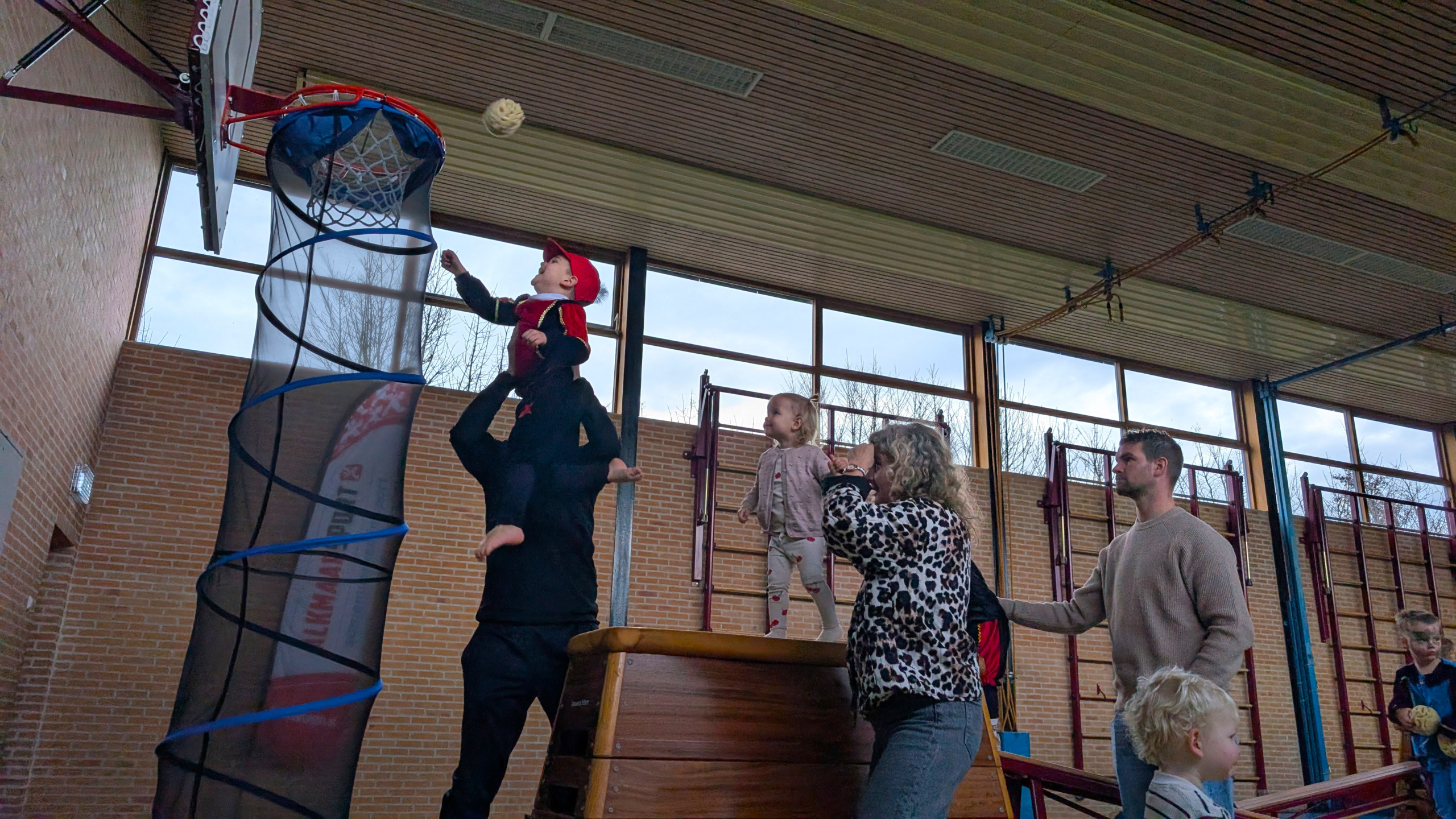 Kinderen en volwassenen in een gymzaal, waarbij een kind op de schouders van een volwassene een bal probeert te gooien in een basket met een lange verenstructuur eronder.
