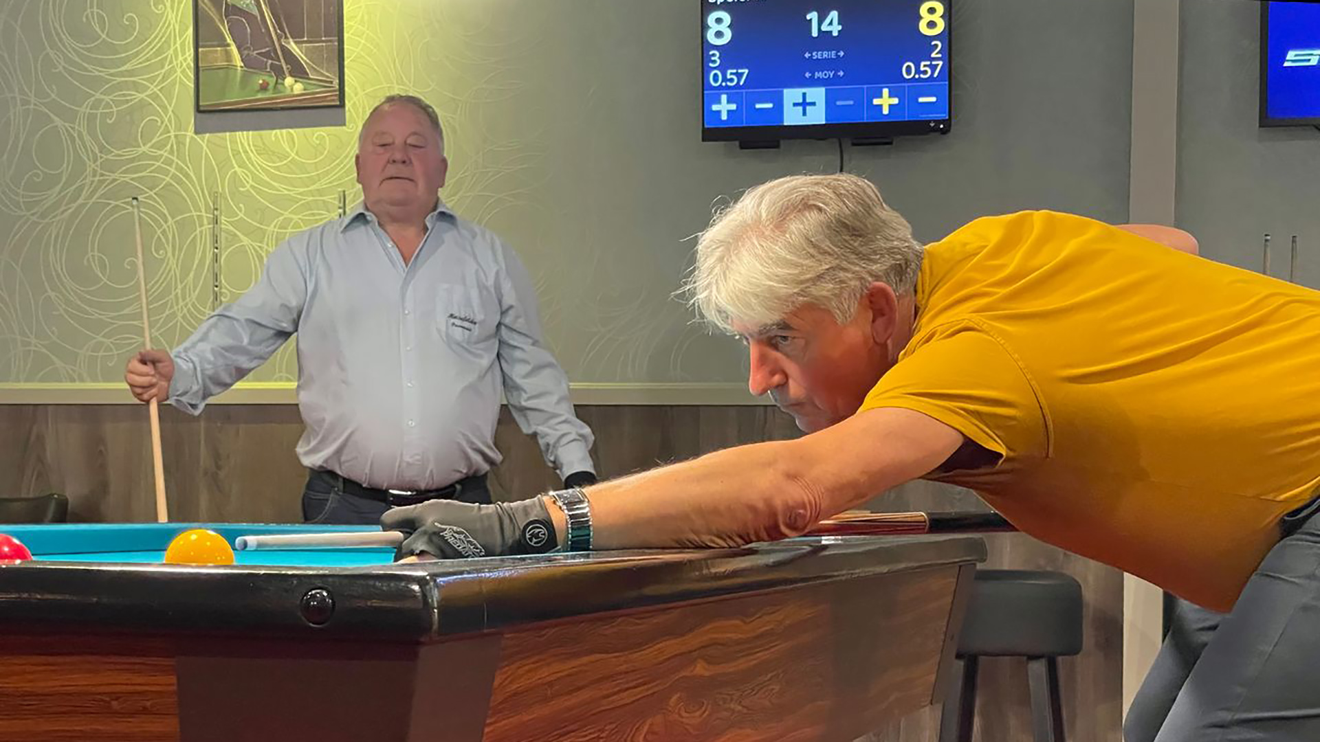 Twee mannen spelen biljart in een zaal, één man richt zich op zijn schot terwijl de ander toekijkt.