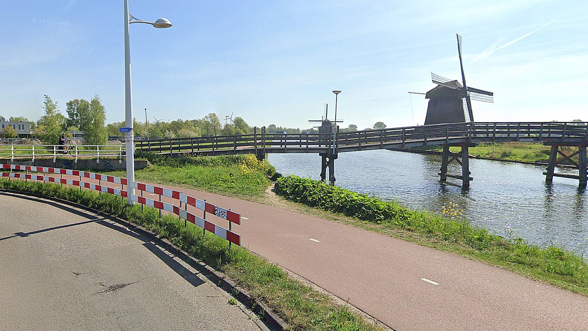 Een houten brug over een waterweg met een traditionele Nederlandse molen op de achtergrond en een fietspad langs de weg.