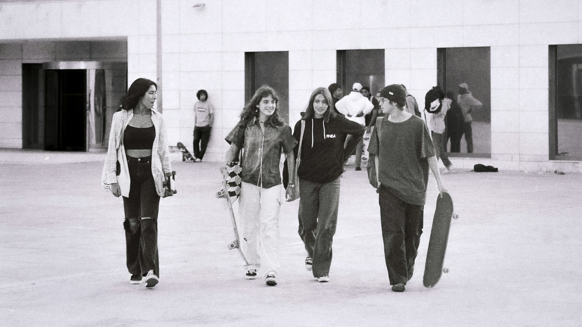 Vier jongeren lopen met skateboards in een open ruimte, lachend en pratend, met een gebouw op de achtergrond.