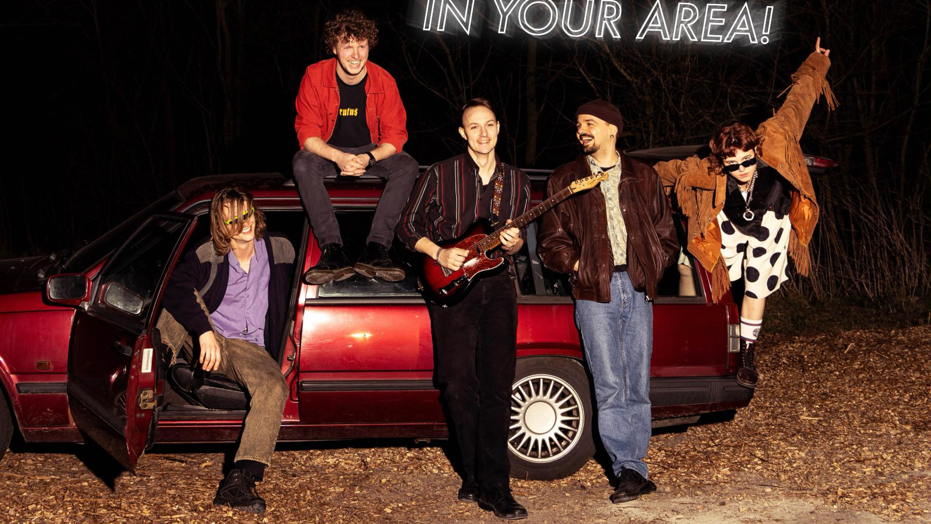 Vijf mensen poseren rond een rode auto; een van hen houdt een gitaar vast, anderen lachen ontspannen.