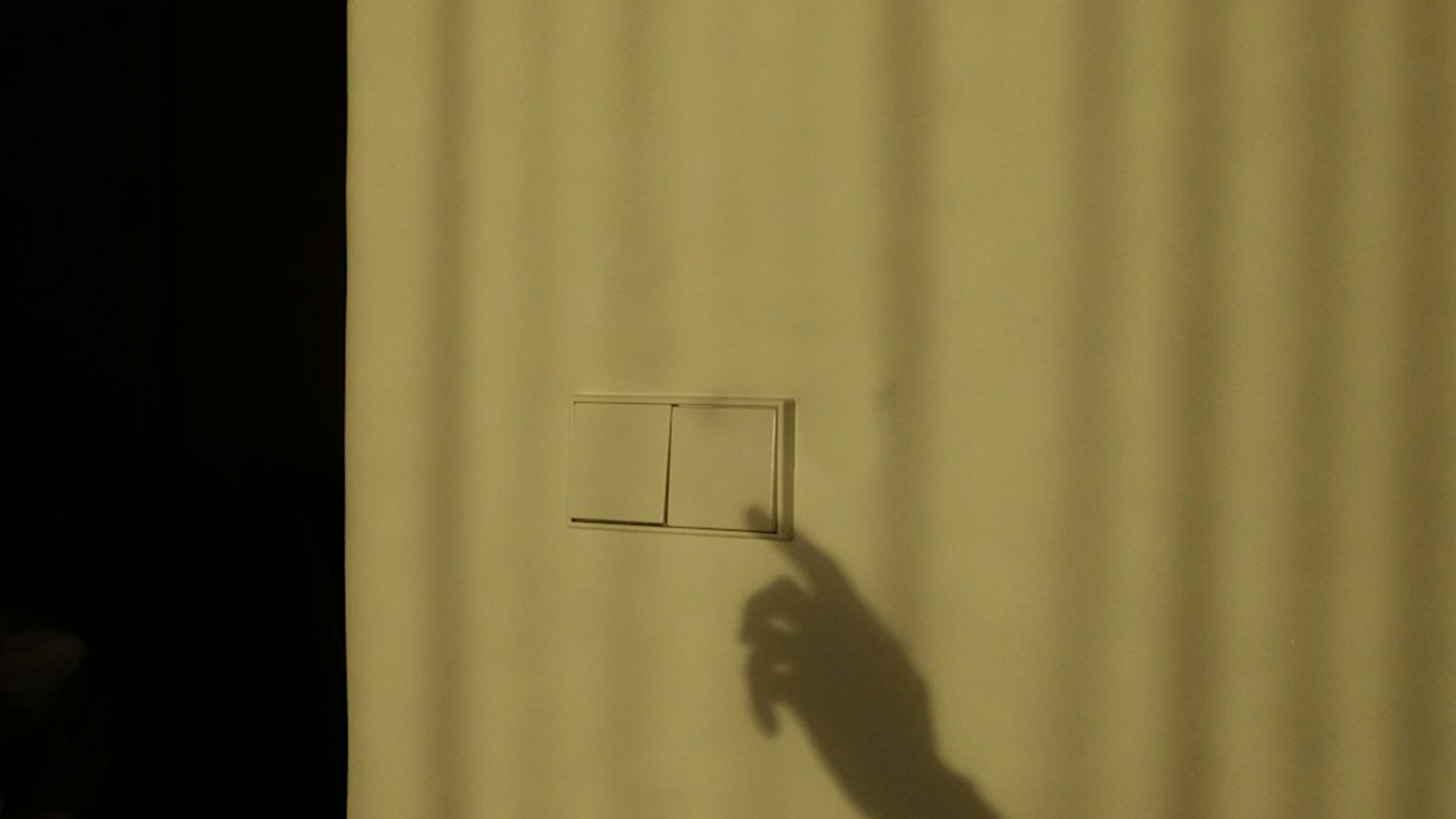 Een schaduw van een hand die naar een lichtschakelaar op een muur wijst, met strepen van zonlicht op de achtergrond.