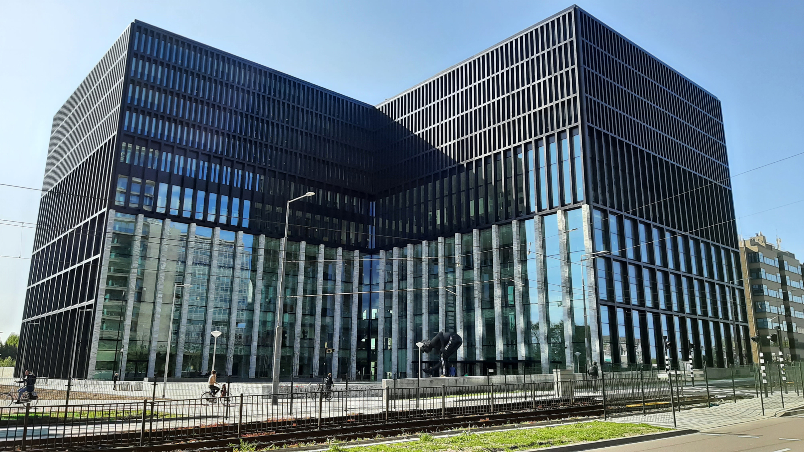 Een modern kantoorgebouw met een glazen gevel en verticale strepen, gelegen aan een straat met fietsers en tramsporen.