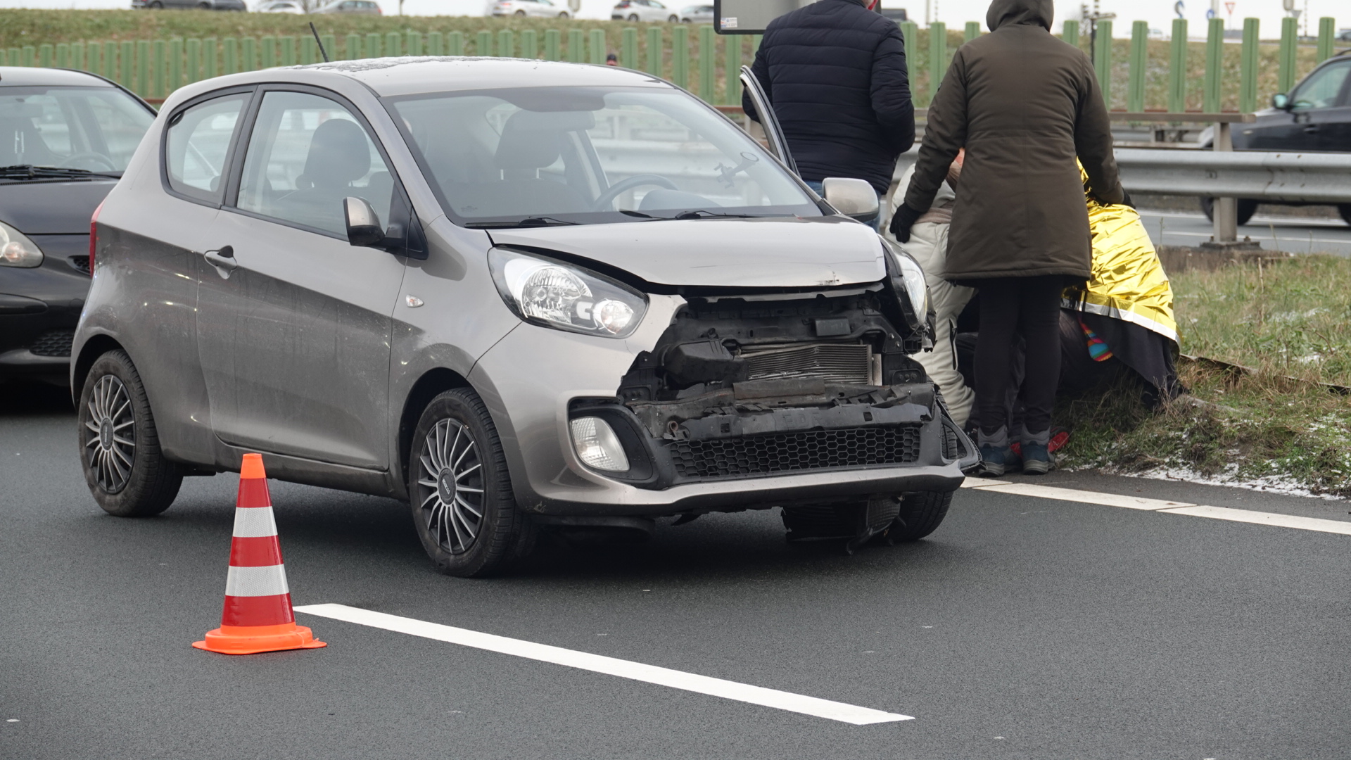 Voorbumperloze zilvergrijze auto op snelweg naast mensen in winterkleding die naast een berm samenkomen, met een verkeerskegel ervoor.