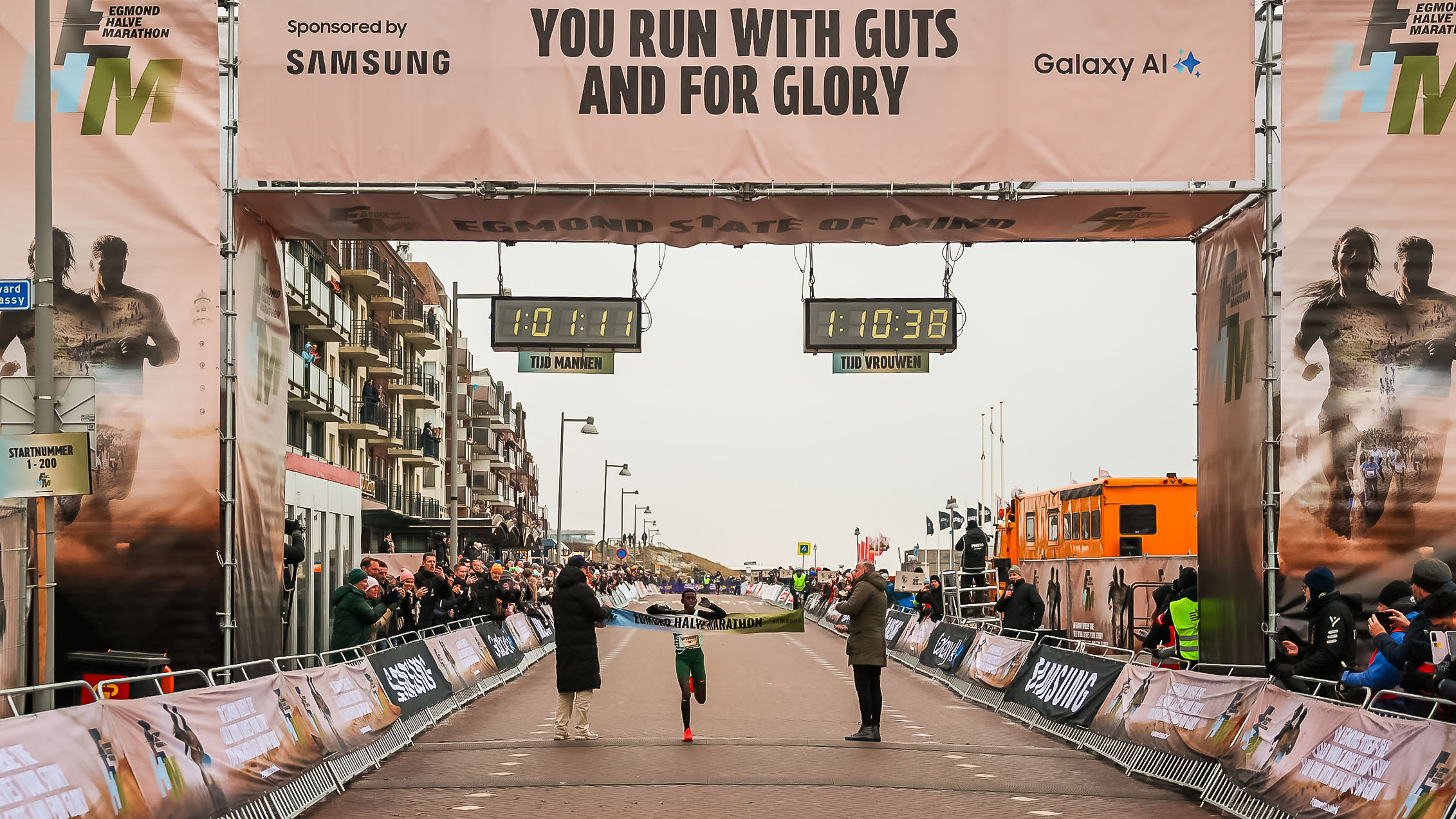 Een hardloper passeert de finish van de Egmond Halve Marathon terwijl toeschouwers applaudisseren, met tijdklokken boven.