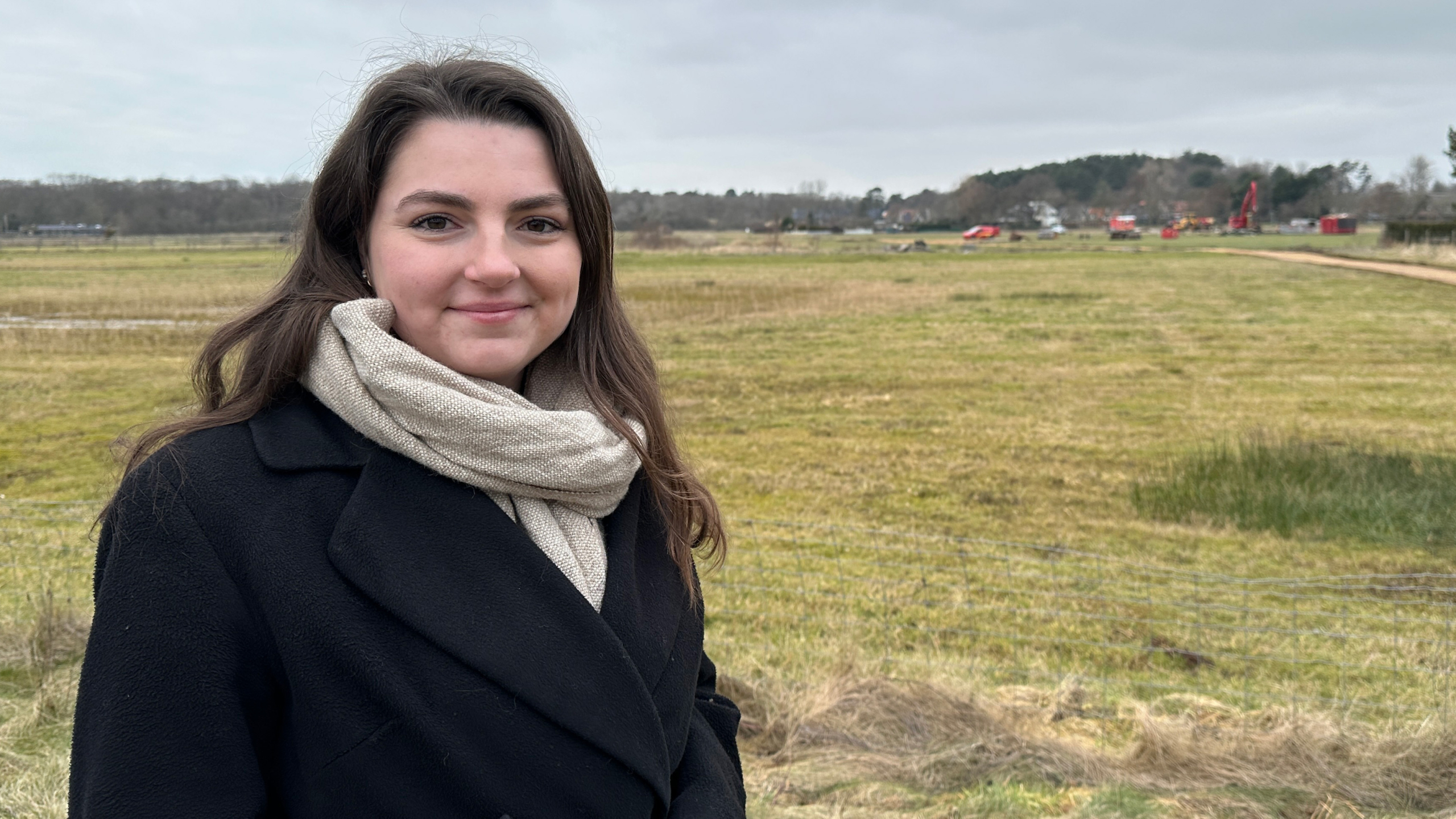 Vrouw met sjaal en zwarte jas staat op een grasveld, met een bouwplaats op de achtergrond.