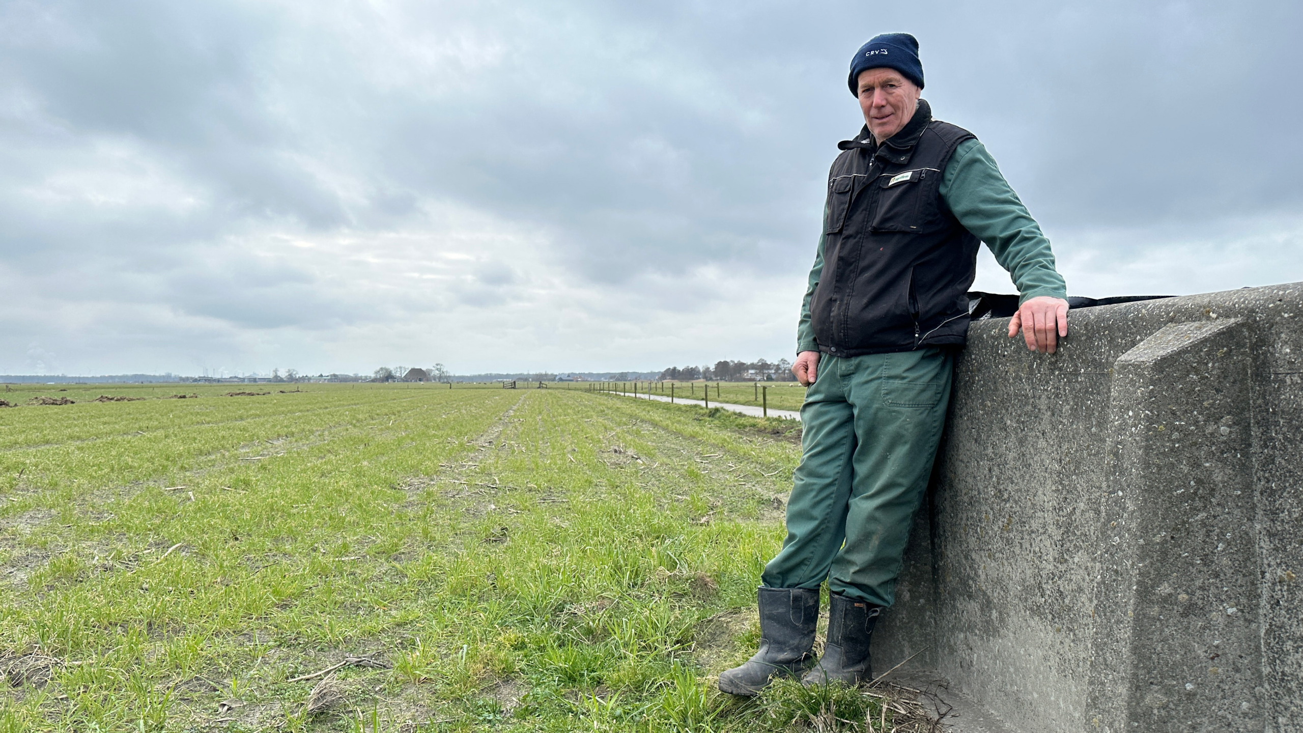 Man in werkkleding leunt tegen een betonnen muur in een weids veld onder een bewolkte lucht.