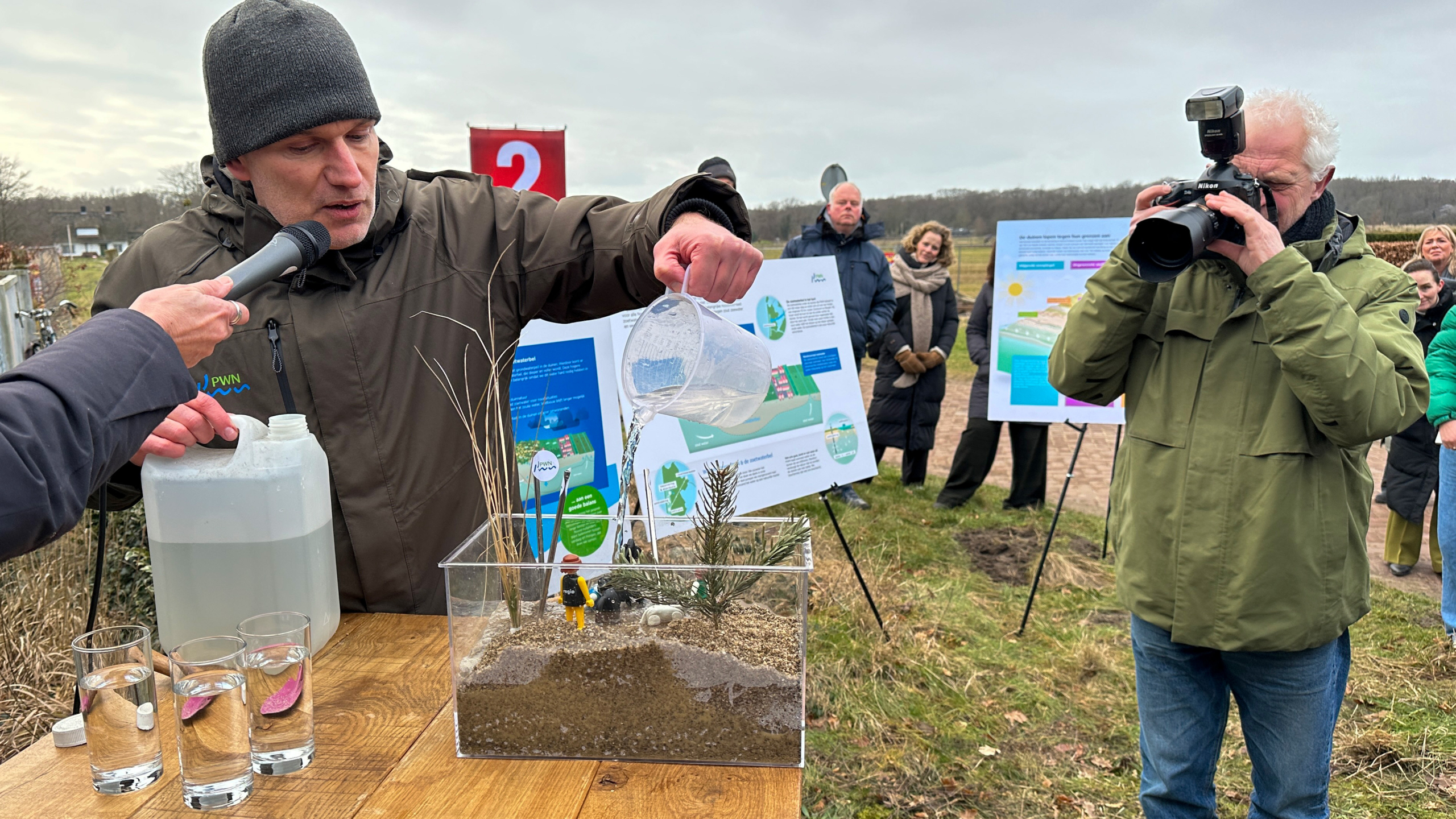 Een man demonstreert het gieten van water in een glazen bak met zand en planten terwijl een fotograaf foto's maakt.