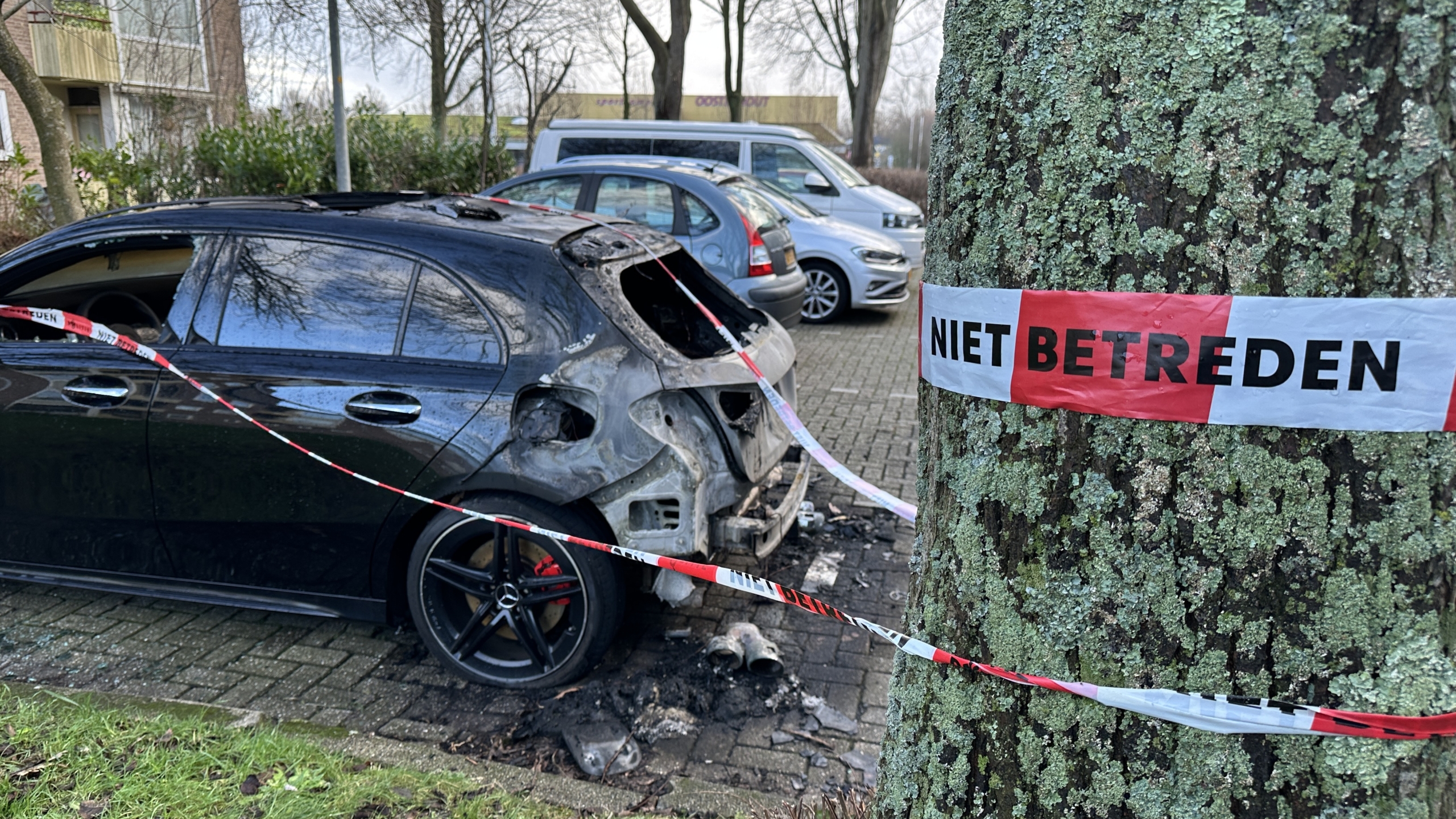 Uitgebrande zwarte auto afgezet met tape waarop "Niet betreden" staat, geparkeerd langs een boom.