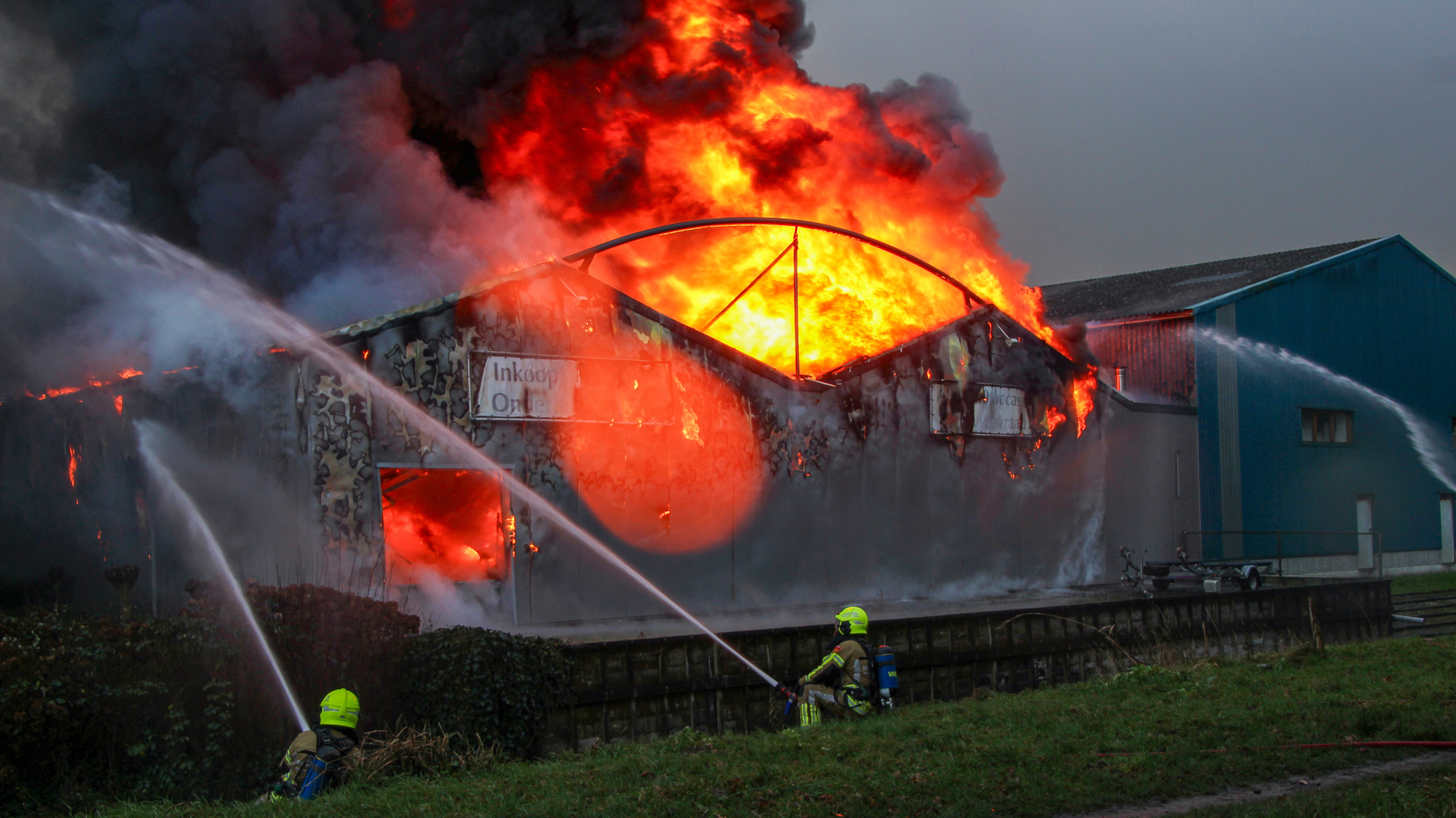 Brandweermannen blussen een intense brand in een loods, waar feloranje vlammen en dikke rookwolken uit het dak slaan.
