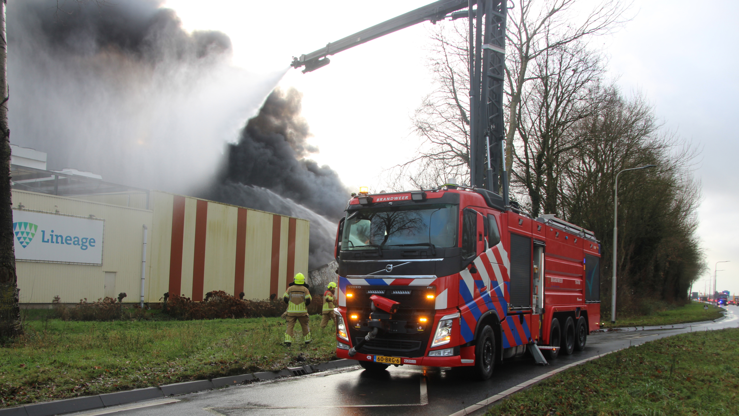 Brandweervoertuig bestrijdt een grote brand met dikke zwarte rook bij een industrieel gebouw met "Lineage" op de gevel.
