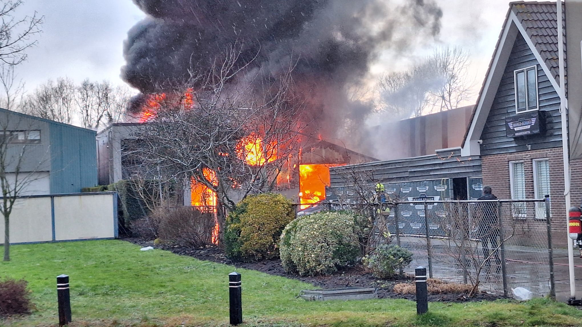 Grote brand met dikke zwarte rook en felle vlammen in een gebouw naast een woonhuis.