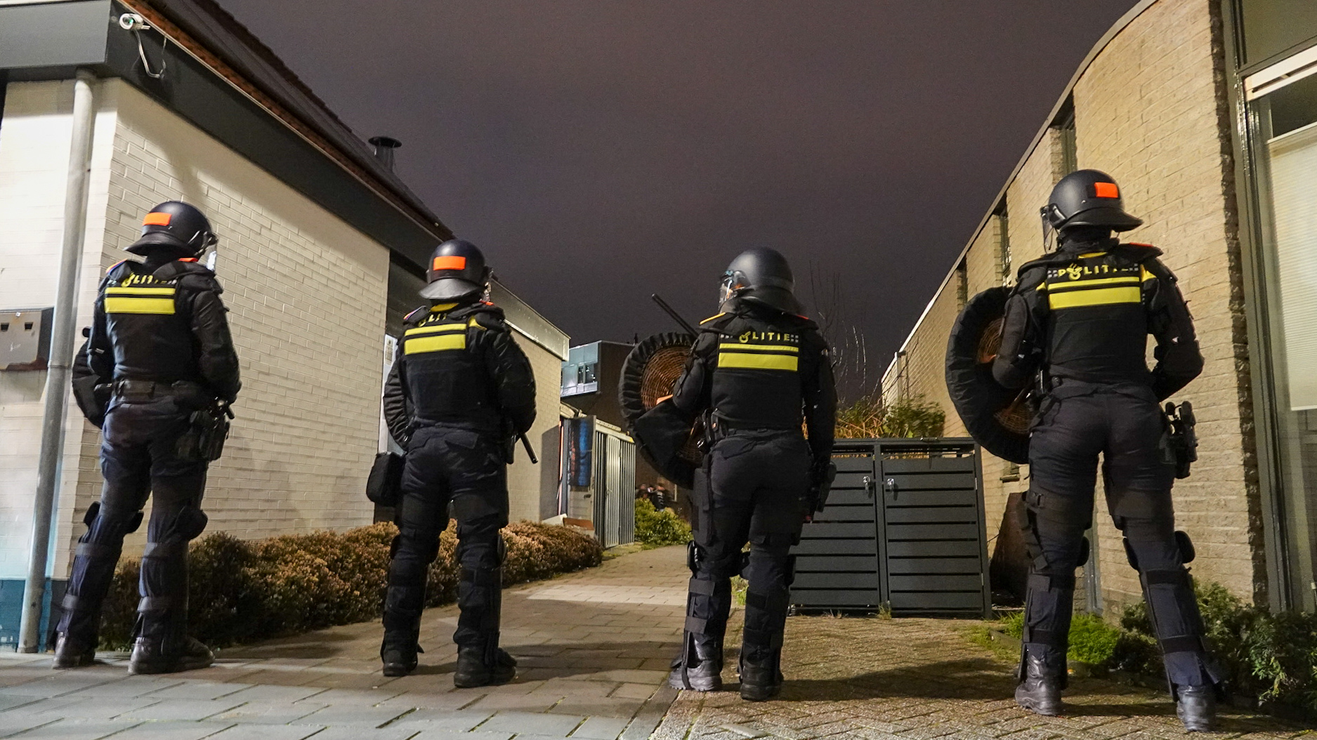 Vier politieagenten in oproeruitrusting staan 's nachts in een smalle straat.