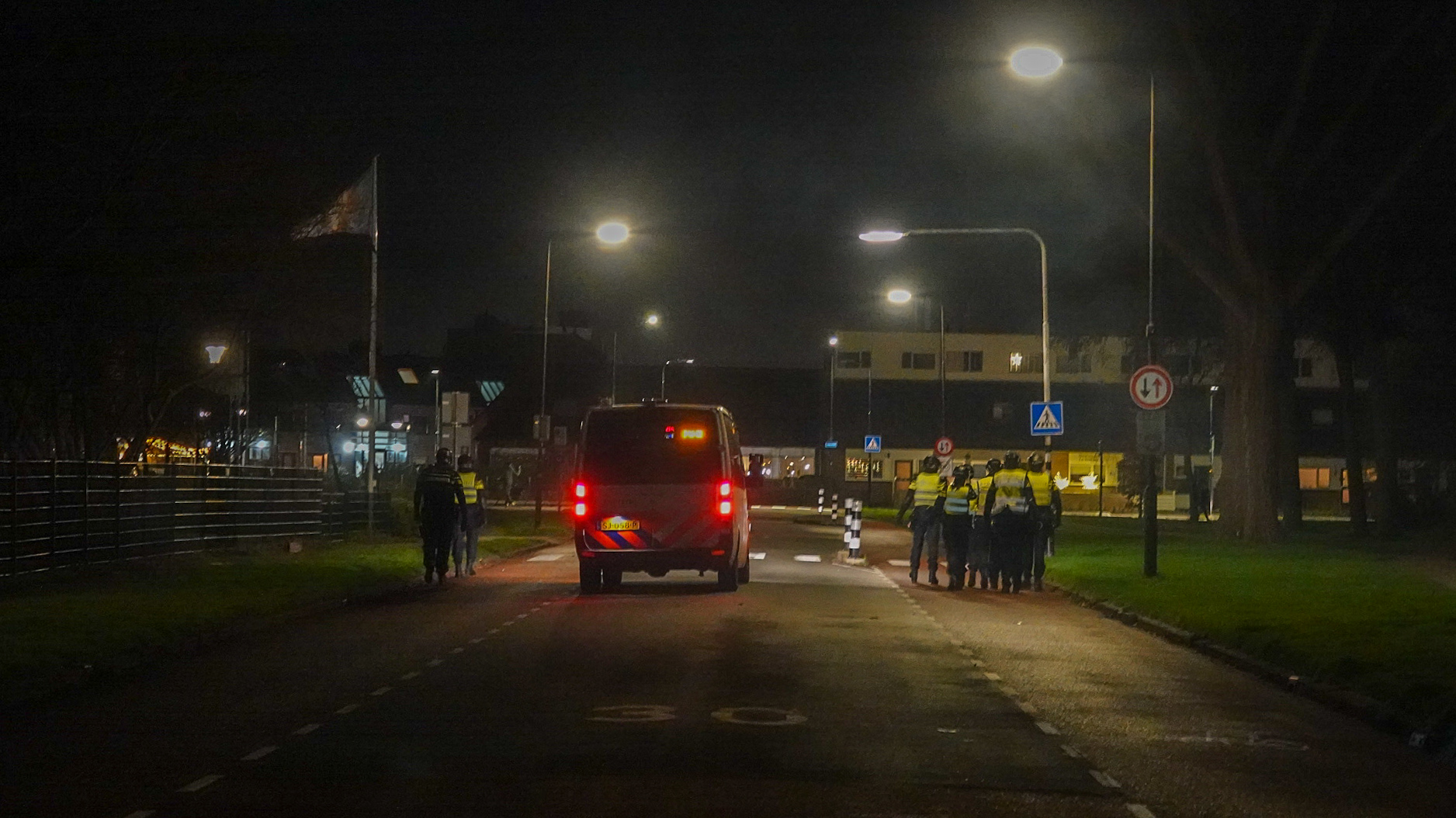 Een politiebusje en agenten staan op een verlichte straat 's nachts, omringd door gebouwen en verkeersborden.