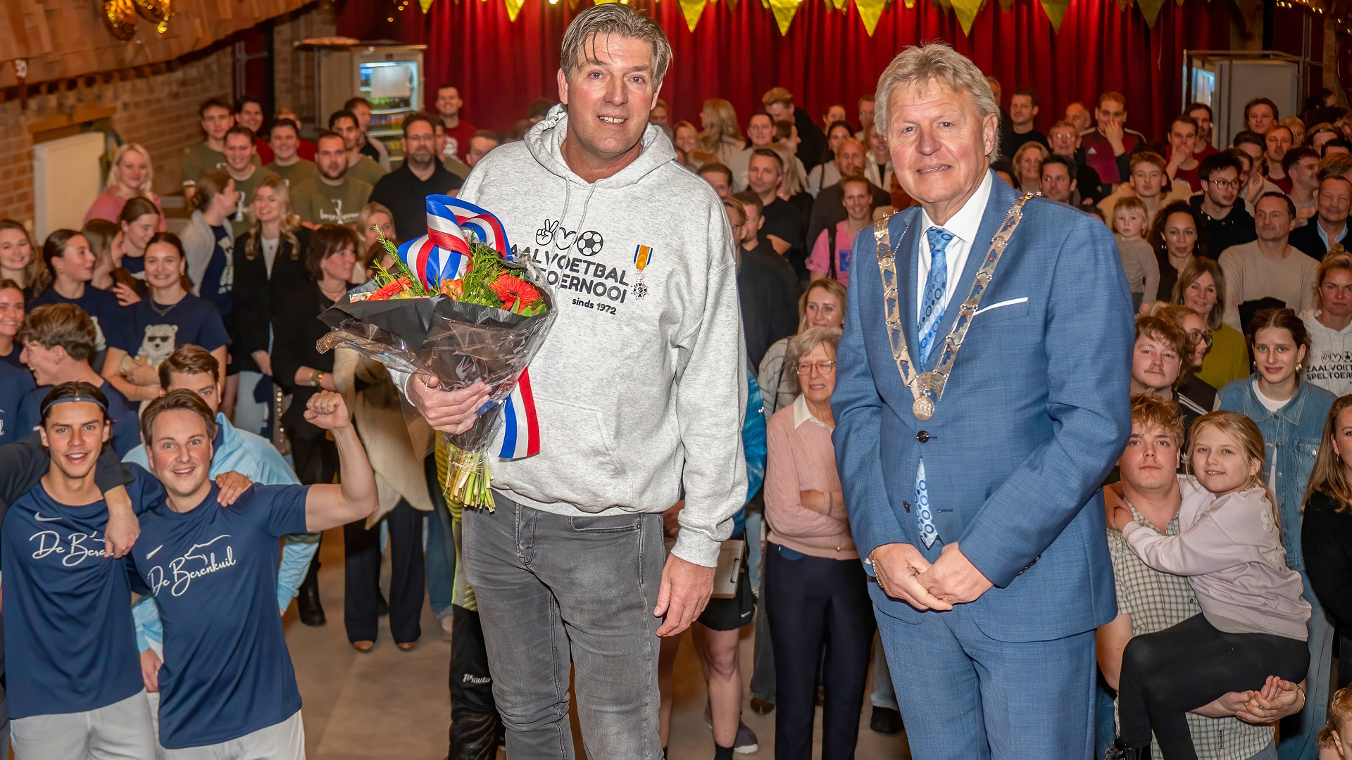 Twee mannen staan voor een groep mensen, één met een bloemenboeket, de ander draagt een ketting, in een feestelijke zaal.