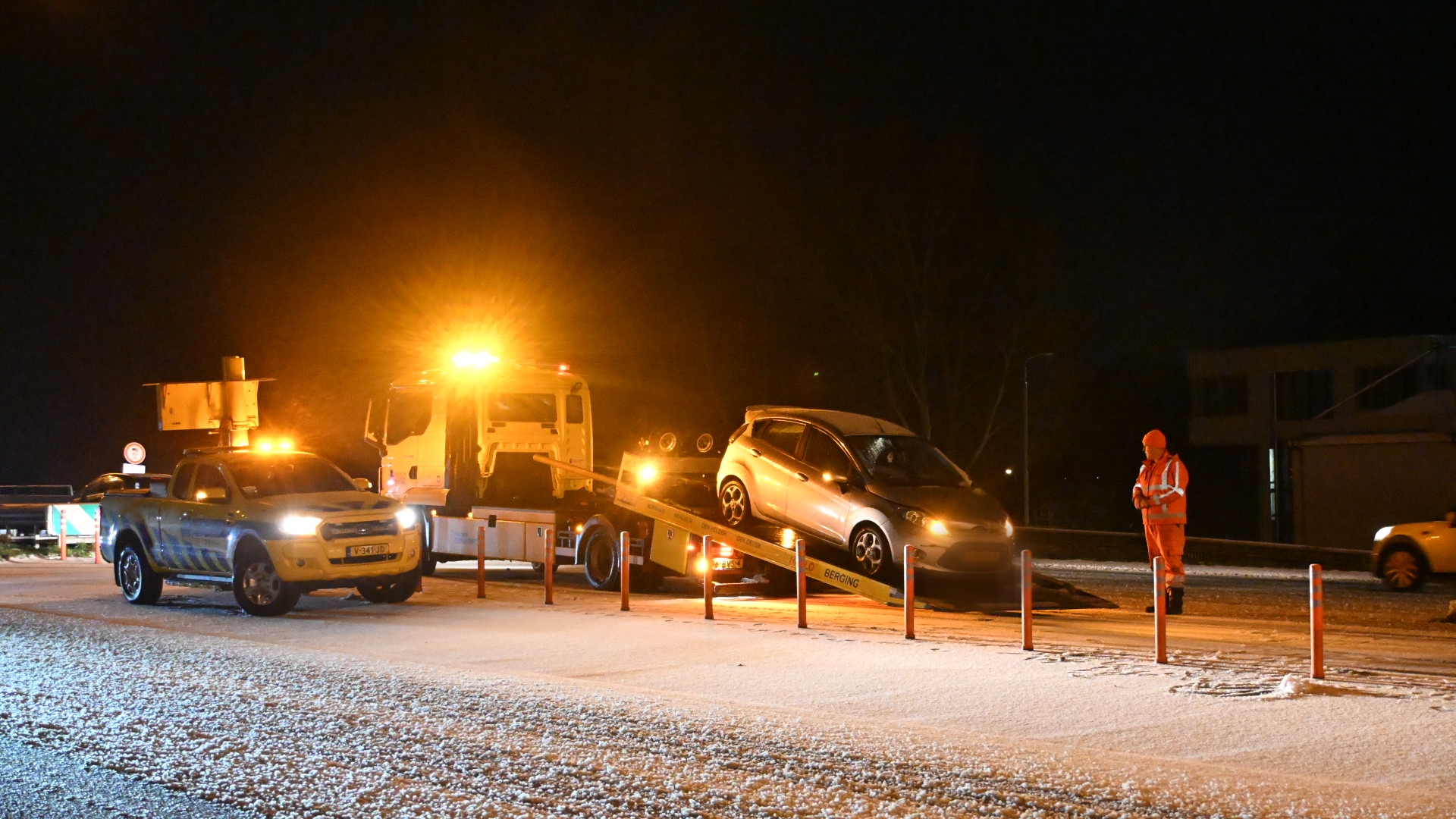 Een auto wordt 's nachts op een takelwagen geladen, begeleid door een persoon in een oranje veiligheidsvest.