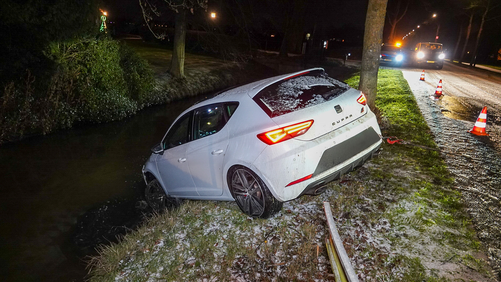 Een witte auto deels in een sloot, met verkeerskegels en een sleepwagen op een besneeuwde weg.