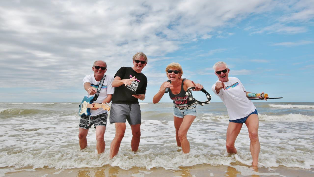Vier lachende mensen op het strand, met instrumenten en in zomerkleding, poseren speels in de branding.
