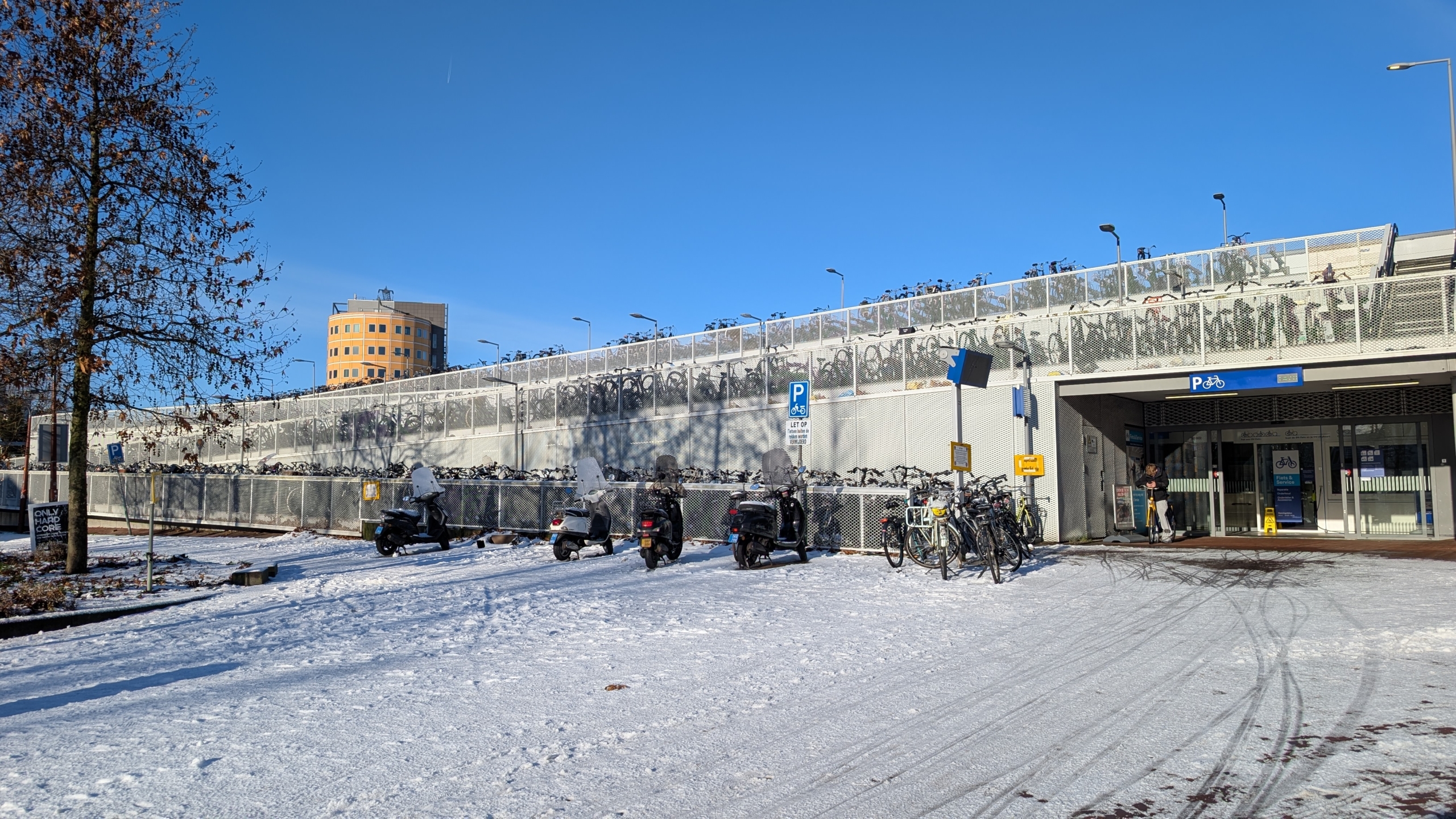 Fietsenstalling met rijen fietsen op meerdere niveaus, op een besneeuwde parkeerplek met een scooter en een gebouw op de achtergrond.