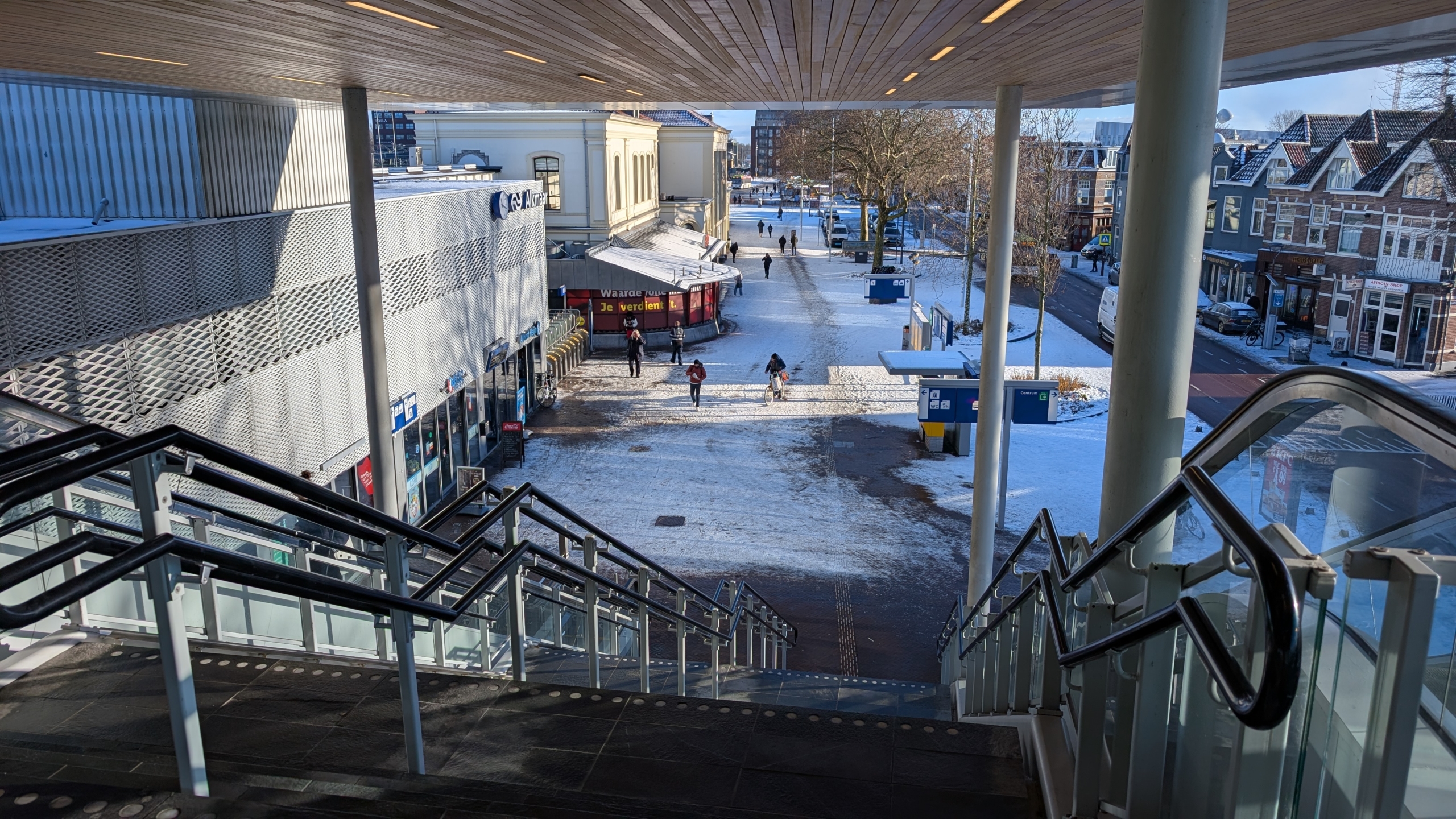 Trap leidt naar besneeuwd plein met winkels, fietsenrek en enkele mensen, omgeven door historische gebouwen en bomen.