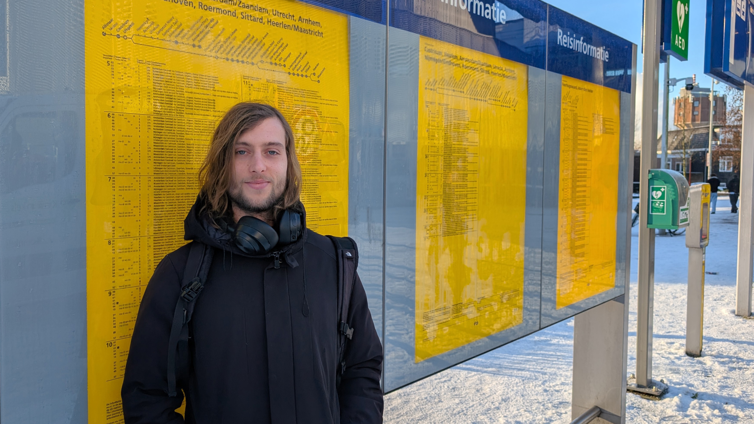 Man met zwarte jas en koptelefoon voor een reisinformatie-bord op een besneeuwd station.