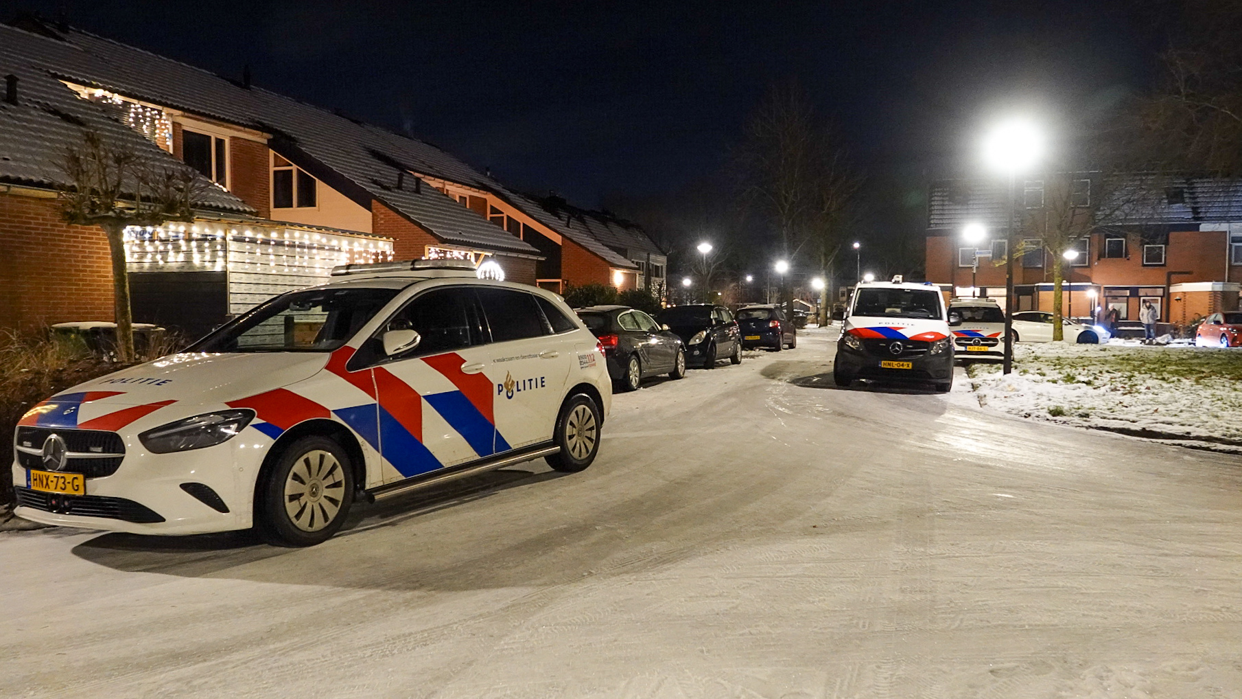 Politieauto's geparkeerd op een besneeuwde woonstraat bij nacht, met huizen versierd met kerstlichtjes.
