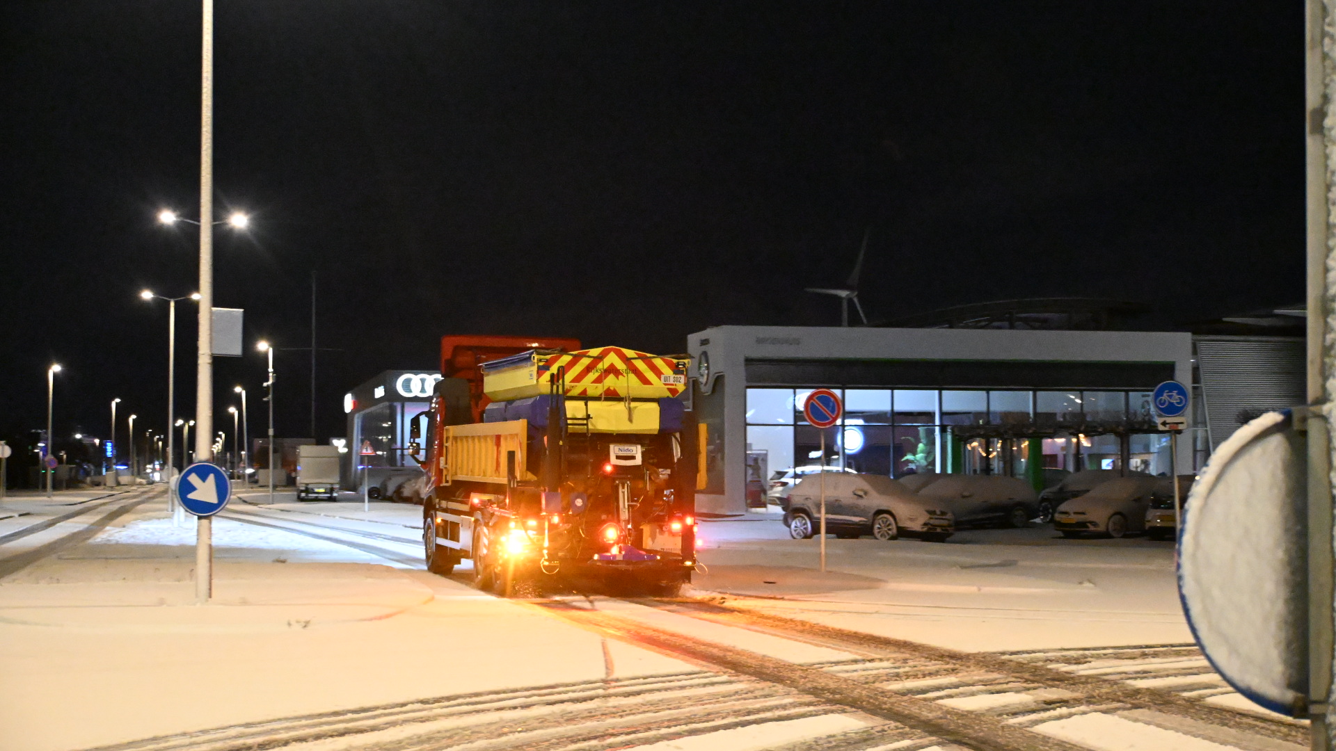 Sneeuwschuiver rijdt 's nachts over besneeuwde weg langs showroom met geparkeerde auto's.