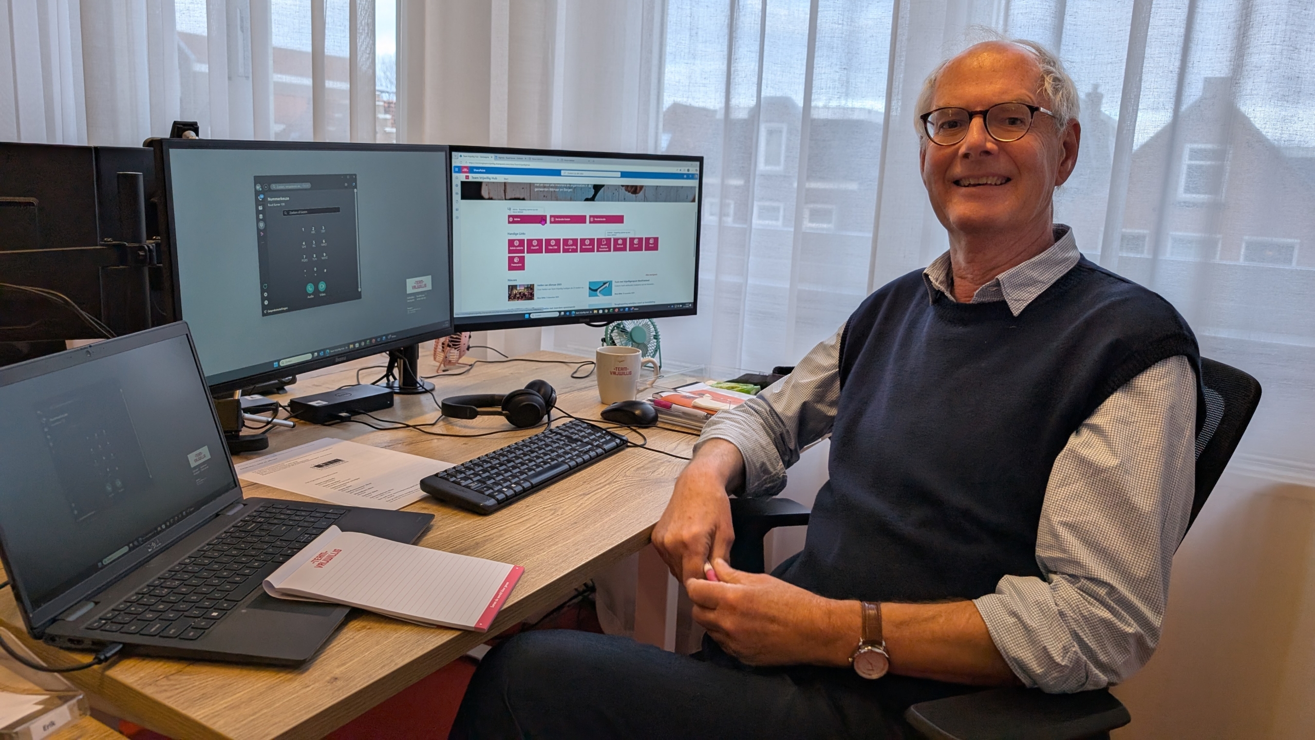 Man achter een bureau met computers en een notitieblok, glimlachend naar de camera in een kantooromgeving.