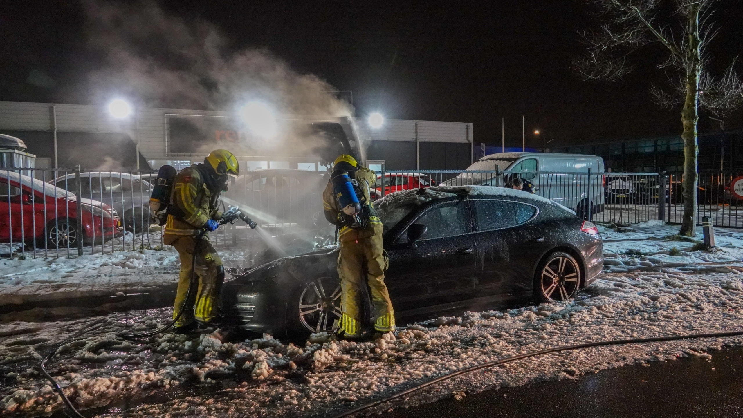 Brandweerlieden blussen 's nachts een brand in een zwarte auto, omringd door schuim en water op de grond.