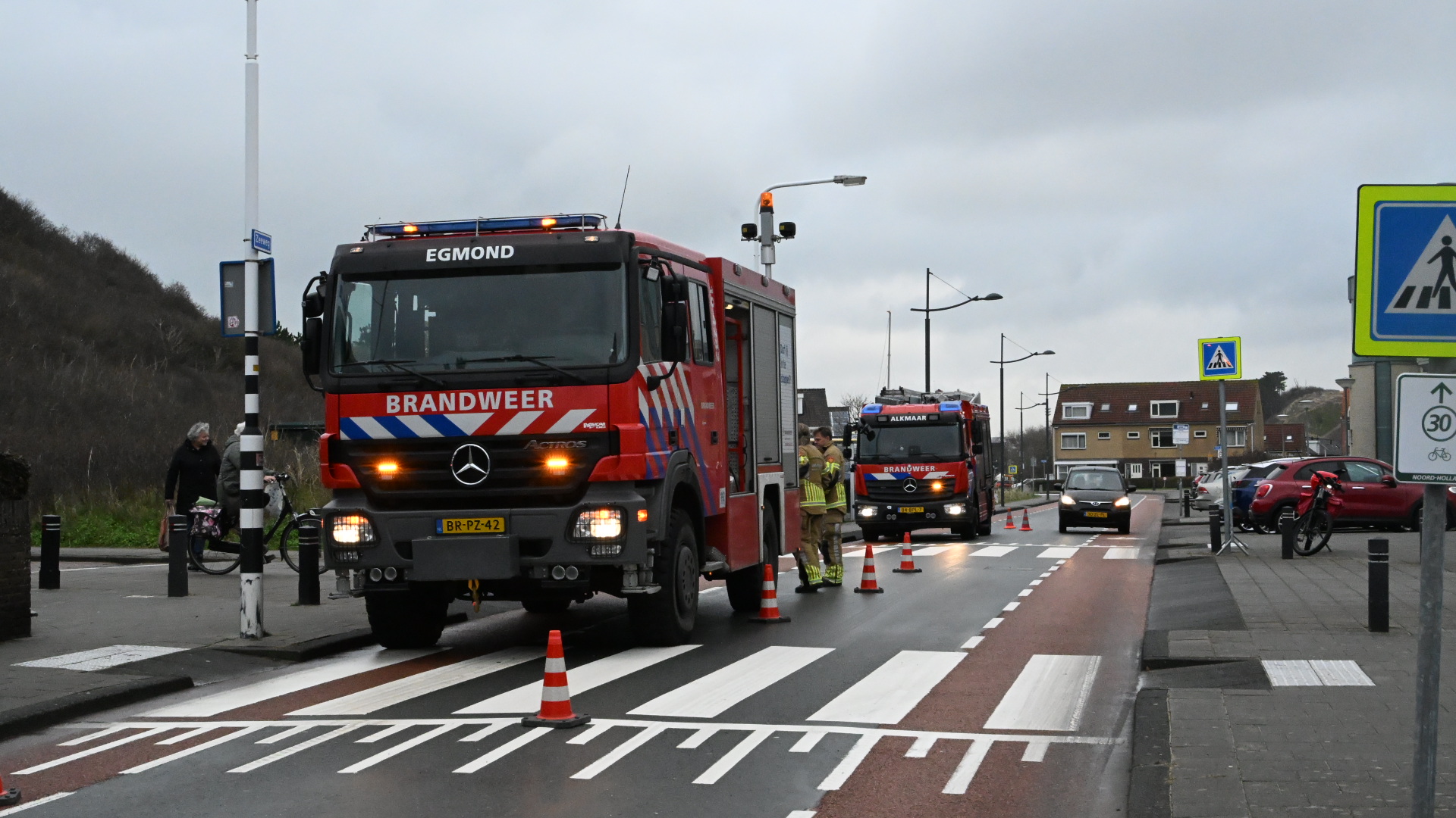 Brandweerwagens naast een zebrapad met verkeerskegels op de weg, omgeven door automobilisten en voetgangers.
