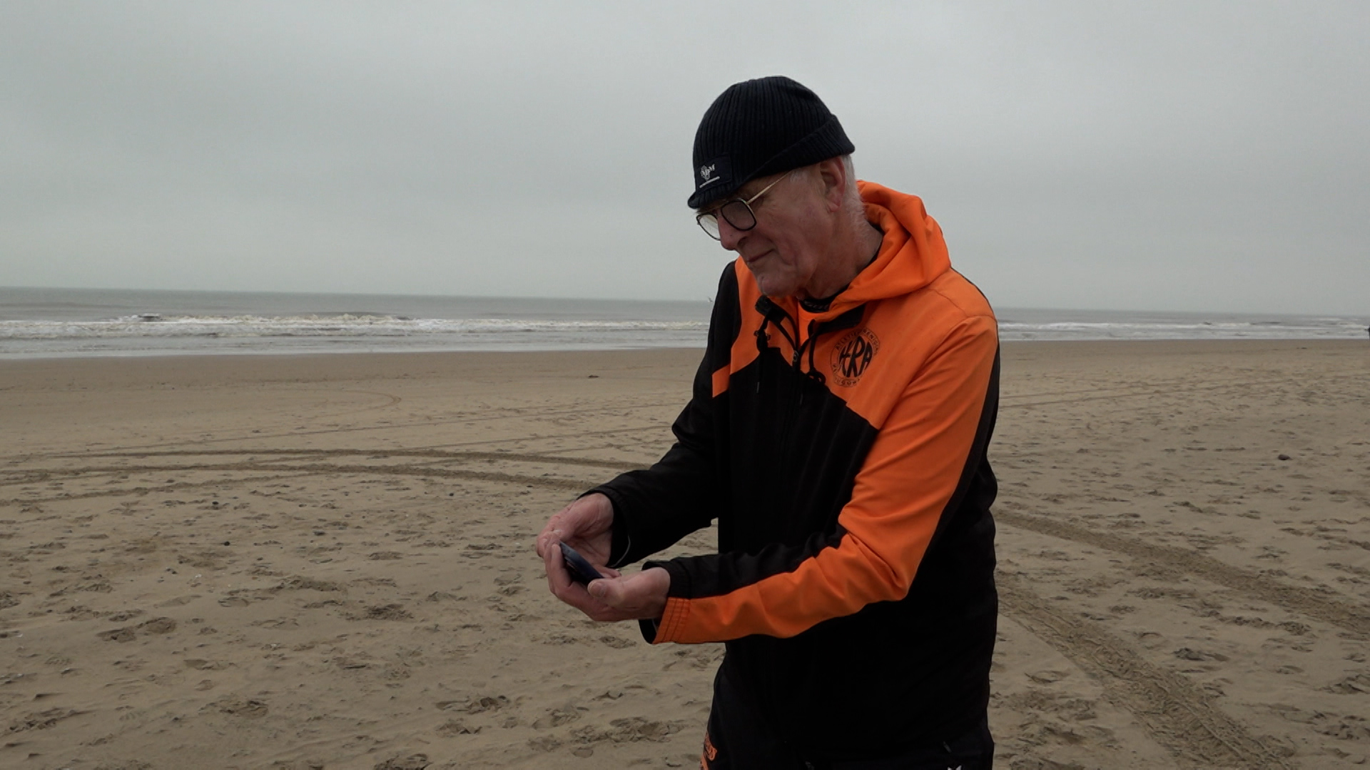 Een man in een oranje en zwarte jas staat op een strand en kijkt naar een voorwerp in zijn handen.