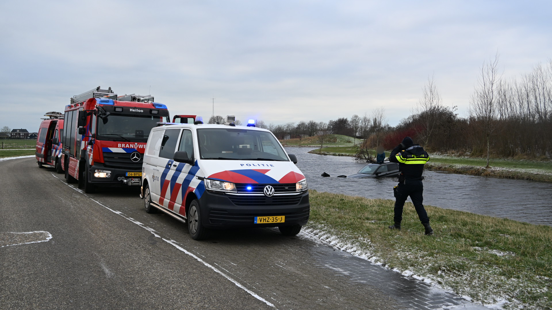 Politie- en brandweerwagens staan aan de kant van de weg bij een half gezonken auto in een sloot, met een agent die toekijkt.