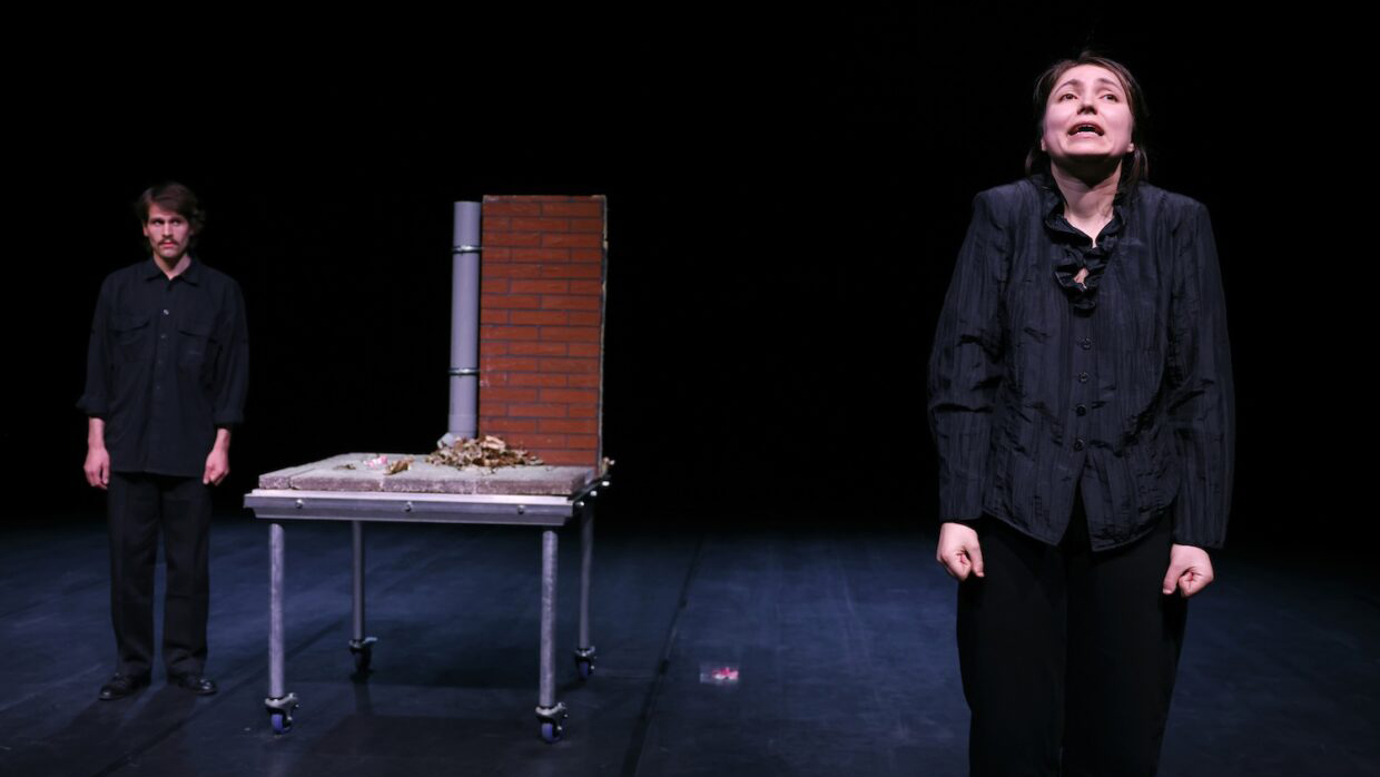 Twee mensen in donkere kleding staan op een toneel met een tafel waarop een bakstenen muur en takken liggen.
