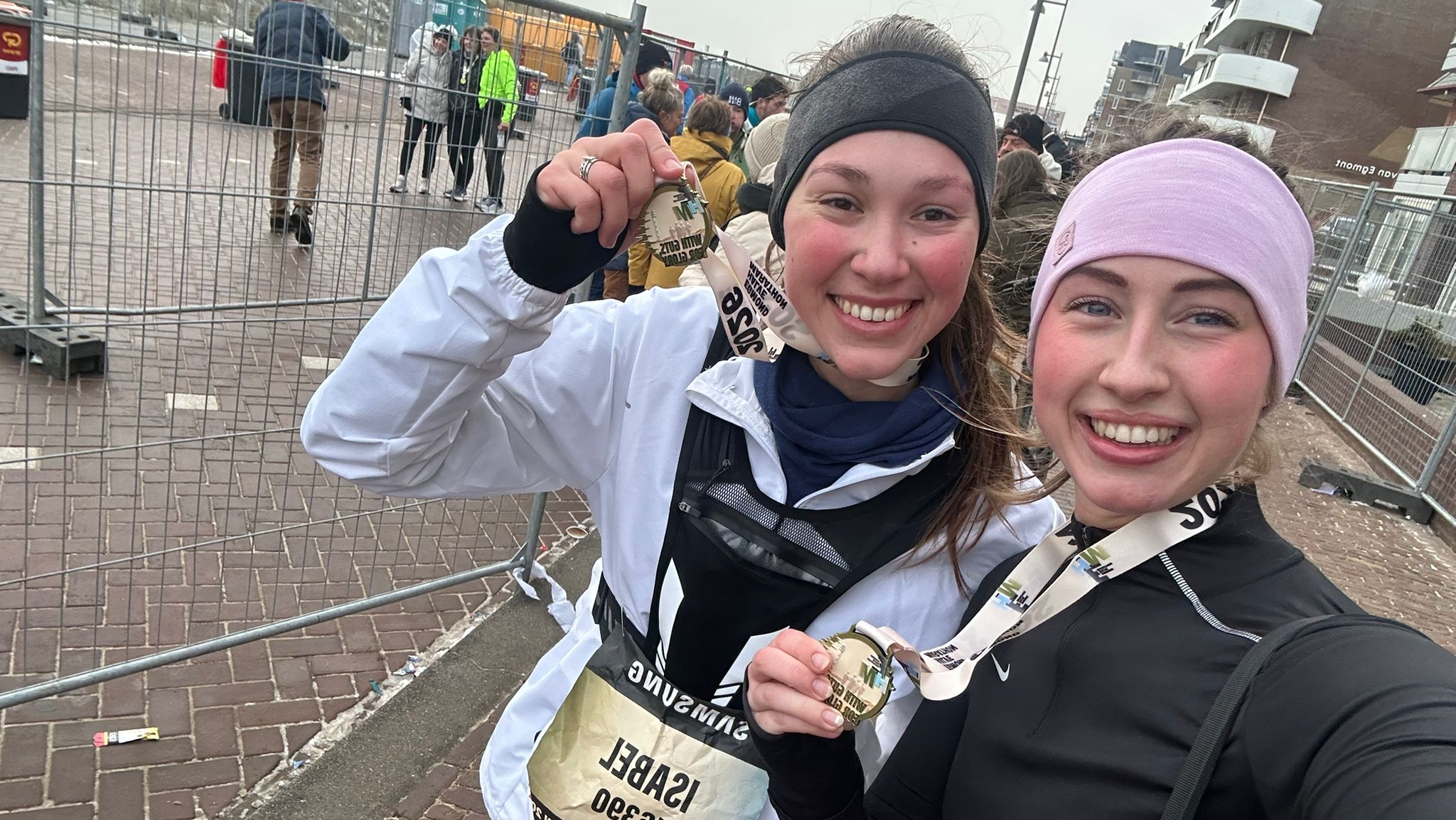 Twee lachende vrouwen met medailles na een marathon, omringd door andere lopers en hekken.