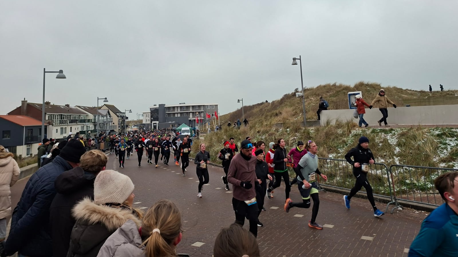 Een groep hardlopers op een besneeuwd pad langs duinen, omringd door toeschouwers en gebouwen op de achtergrond.