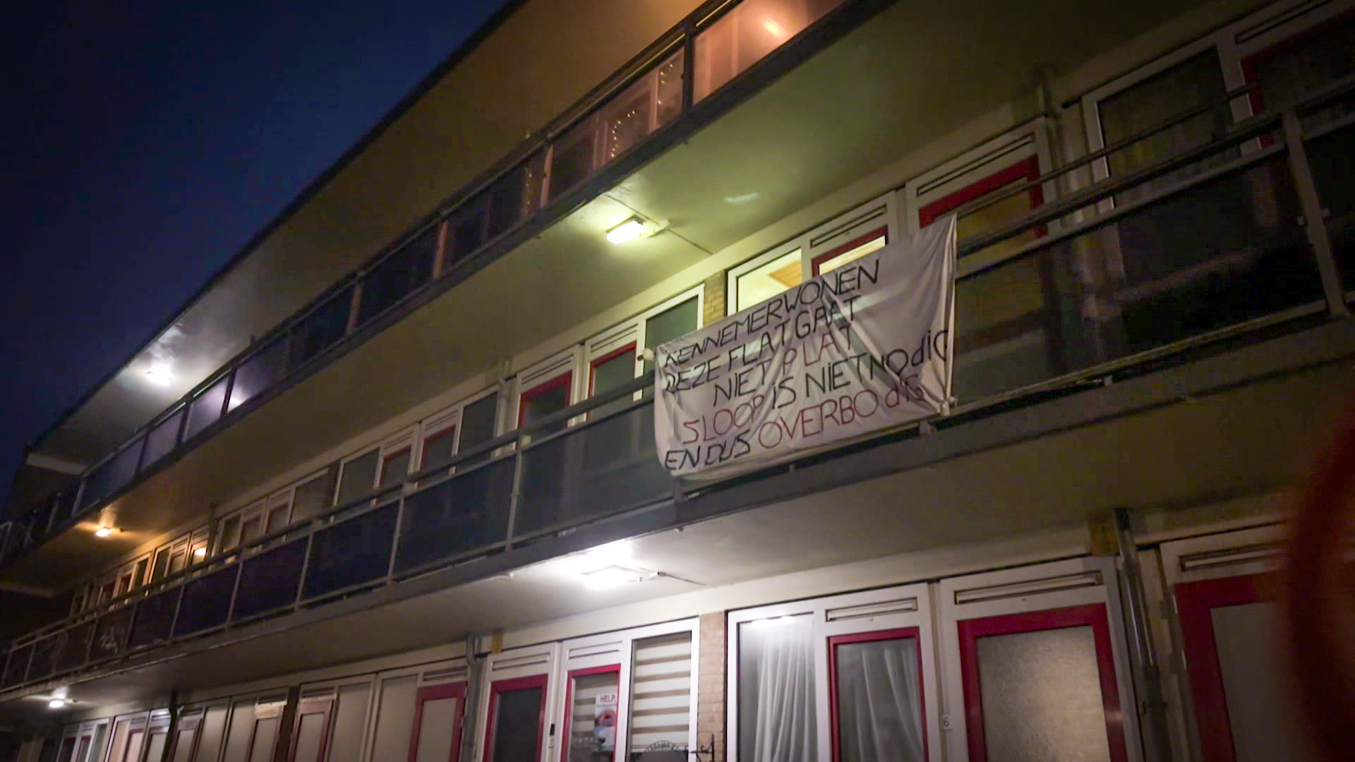 Flatgebouw bij nacht met een spandoek aan de balustrade met protesttekst over sloopplannen.