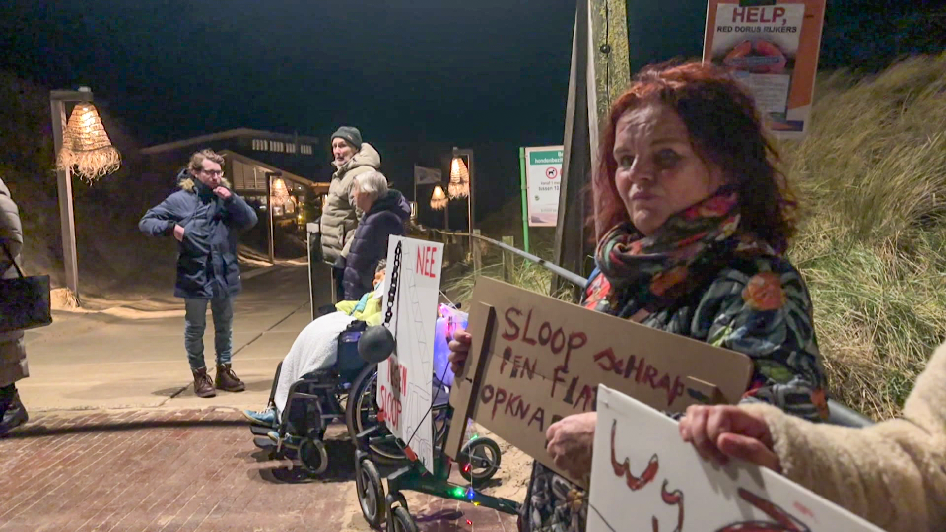 Mensen protesteren 's avonds buiten met borden waarop teksten staan, zoals "Sloop" en "Nee".