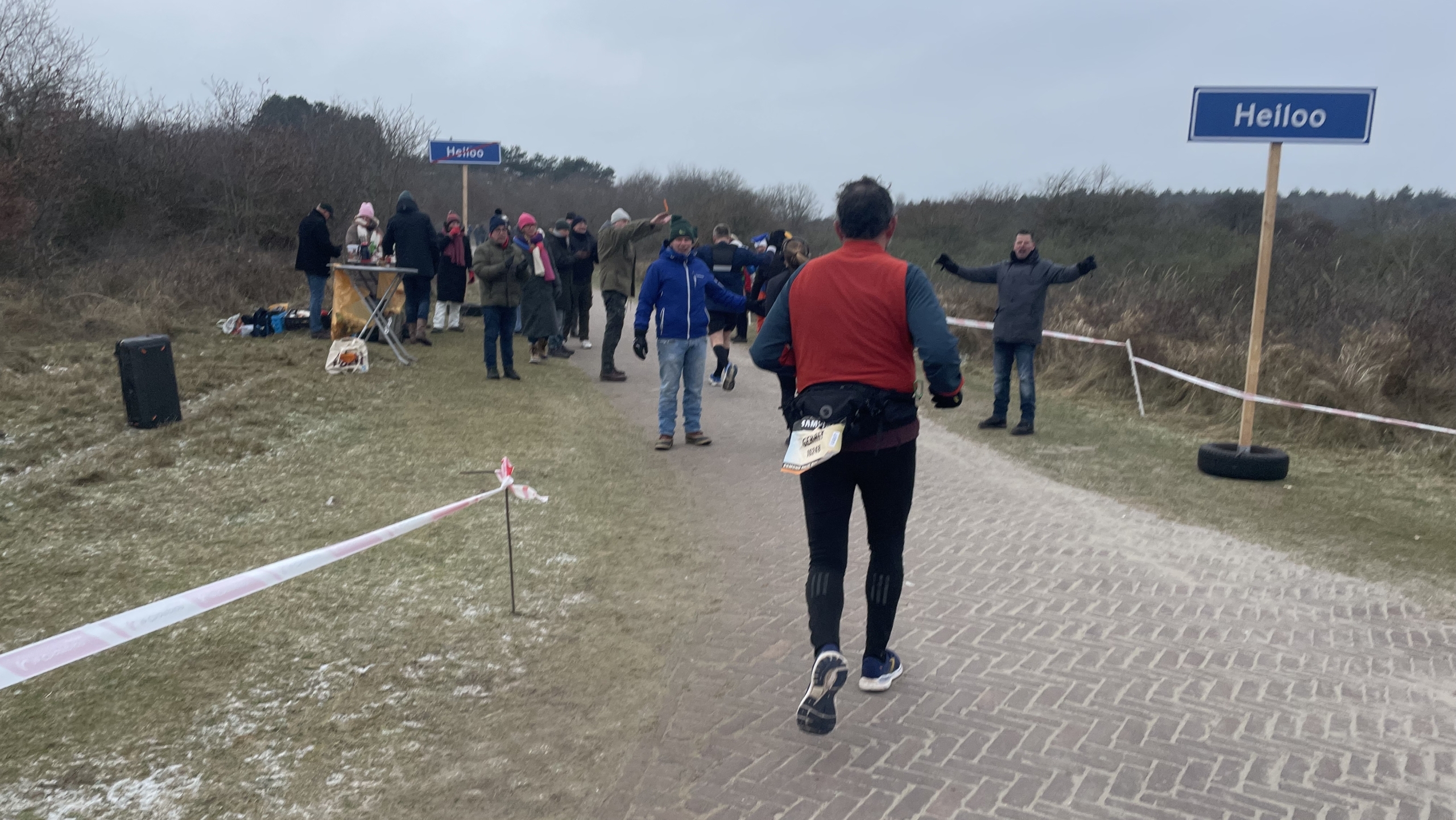 Hardloper nadert een groep toeschouwers langs een pad, met een straatbord "Heiloo" en wat mensen bij een tafel.