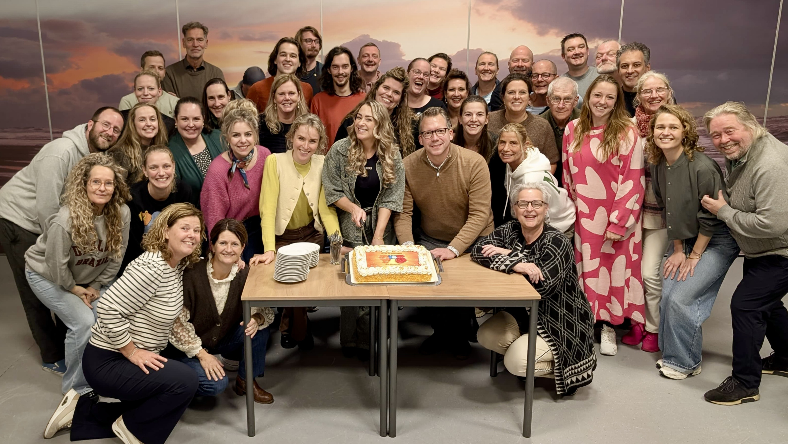 Een grote groep mensen poseert lachend rond een taart op een tafel, klaar om te snijden, met een geschilderde zonsondergang op de achtergrond.