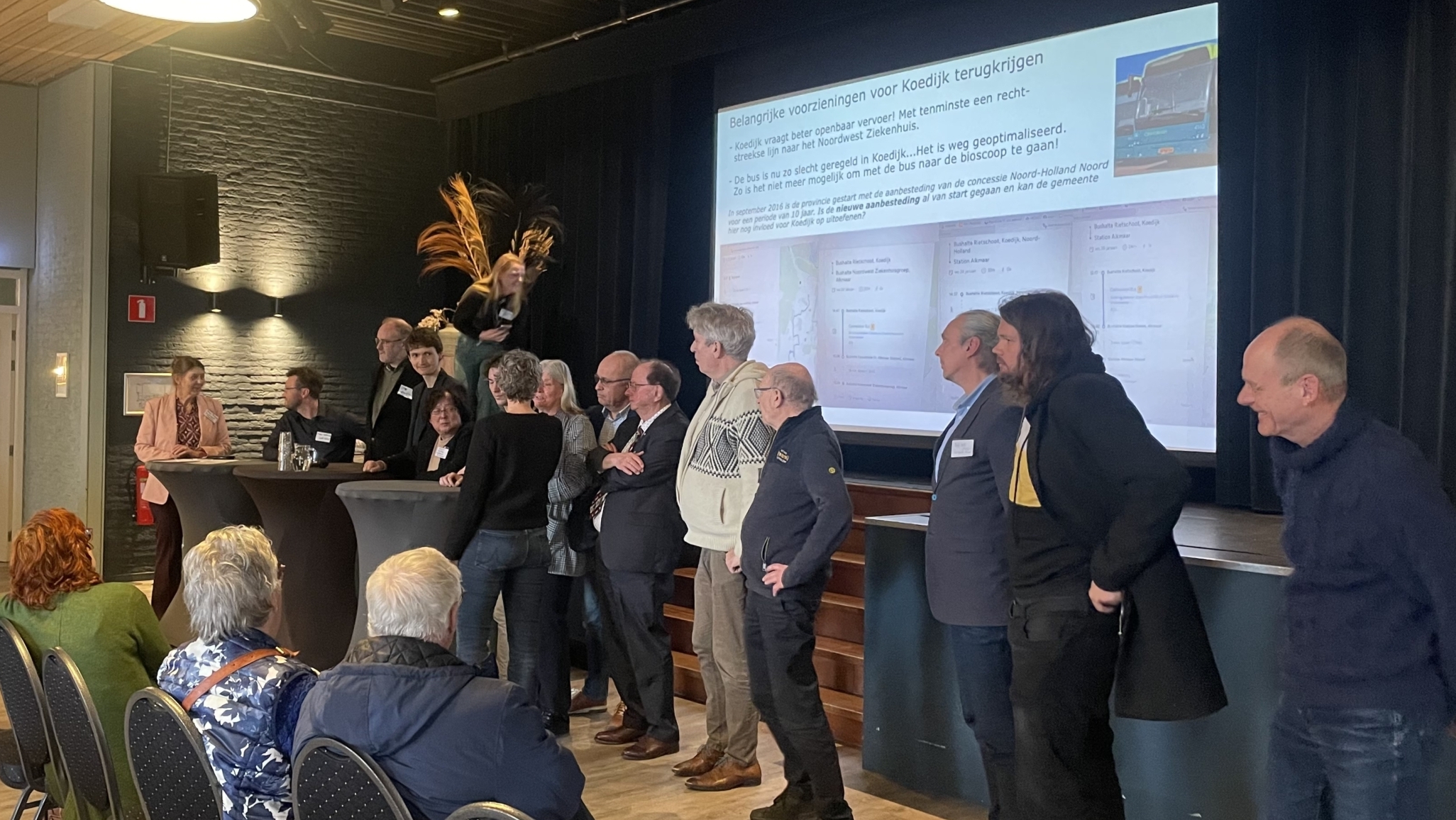Een groep mensen staat op een podium voor een projectiescherm tijdens een presentatie in een zaal met toeschouwers.