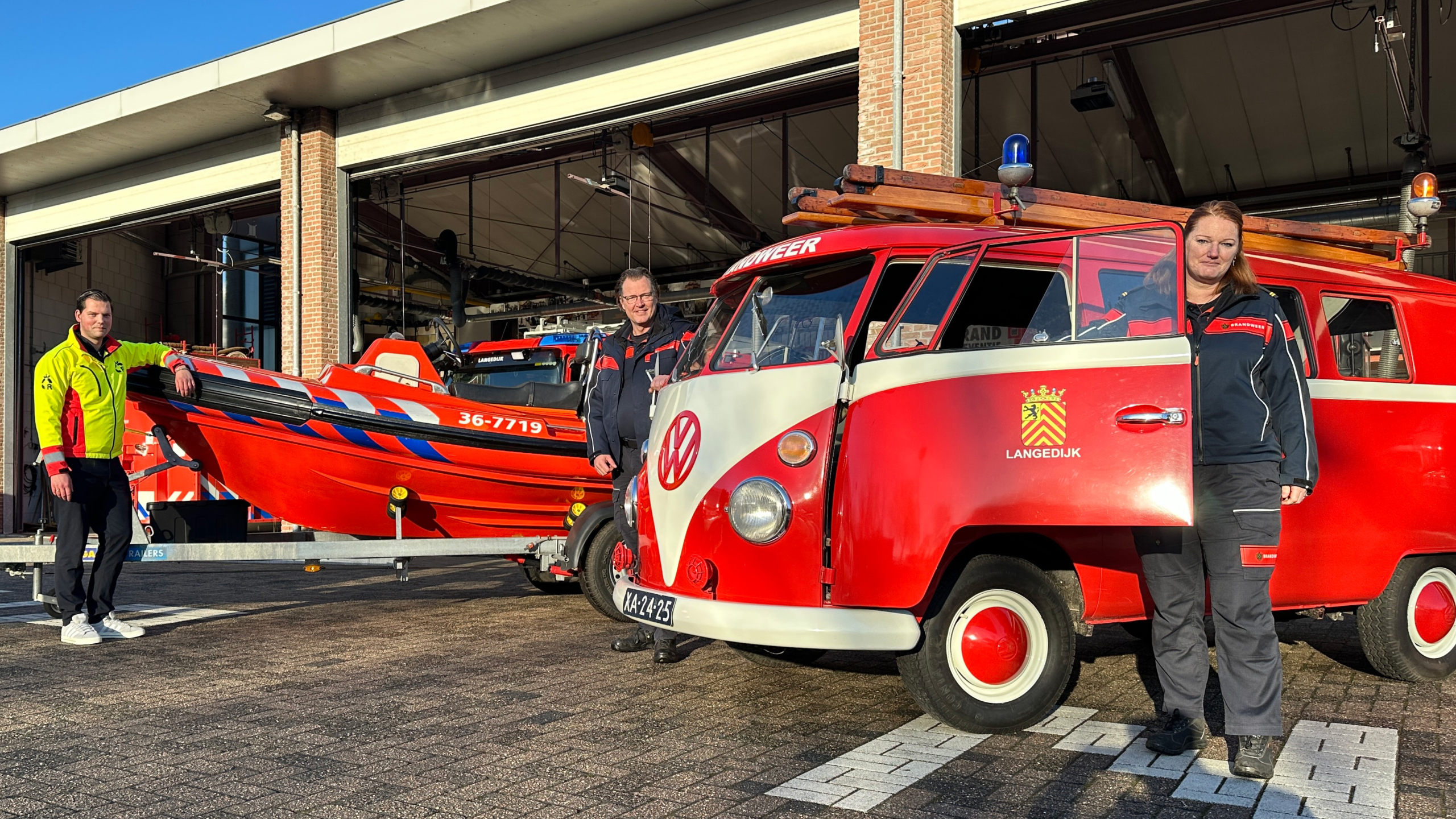Drie brandweerlieden poseren buiten een brandweerkazerne met een rode Volkswagen bus en een reddingsboot.