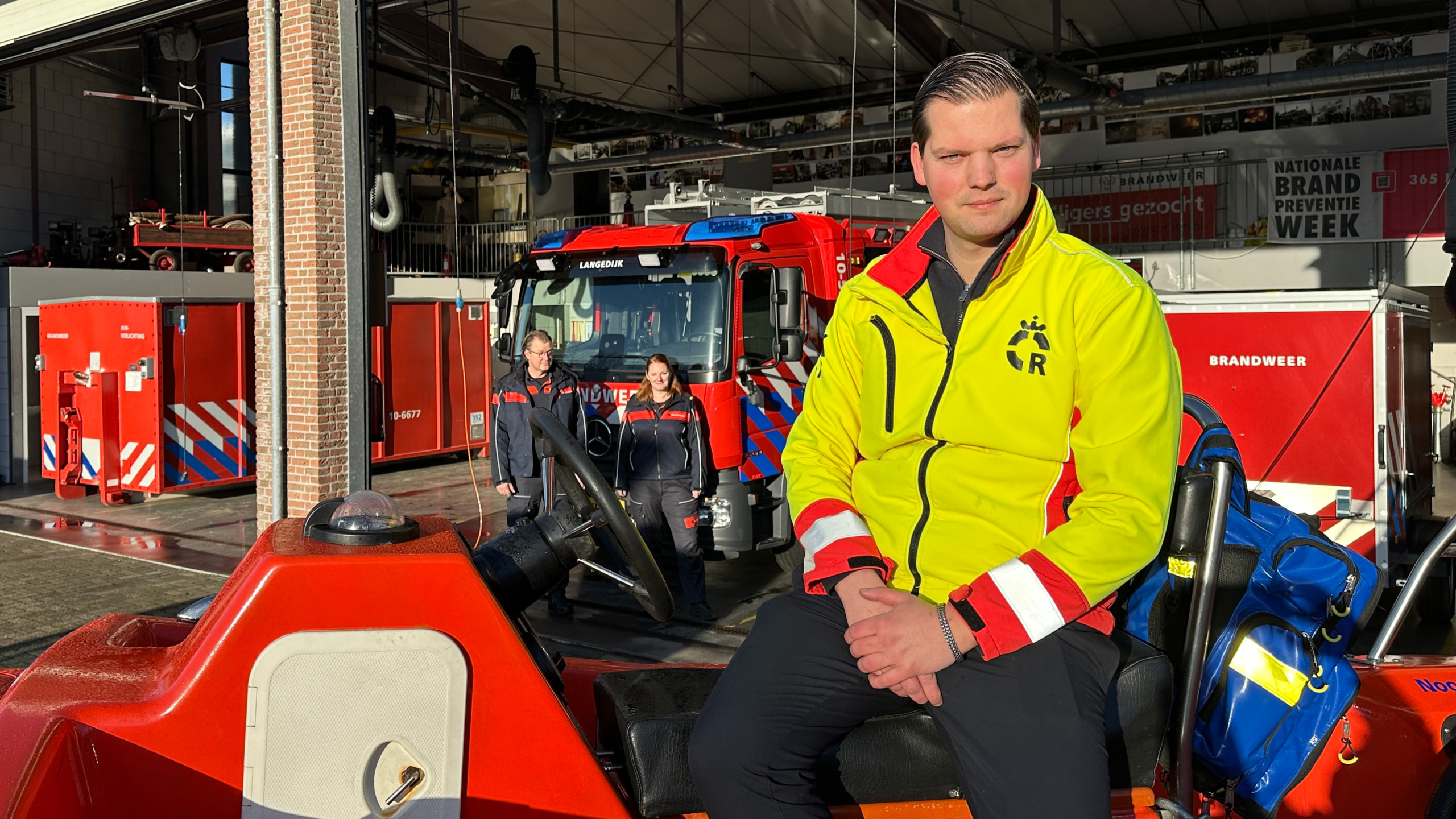 Een man in een gele jas zit op een voertuig voor een brandweerkazerne met twee brandweerlieden op de achtergrond.