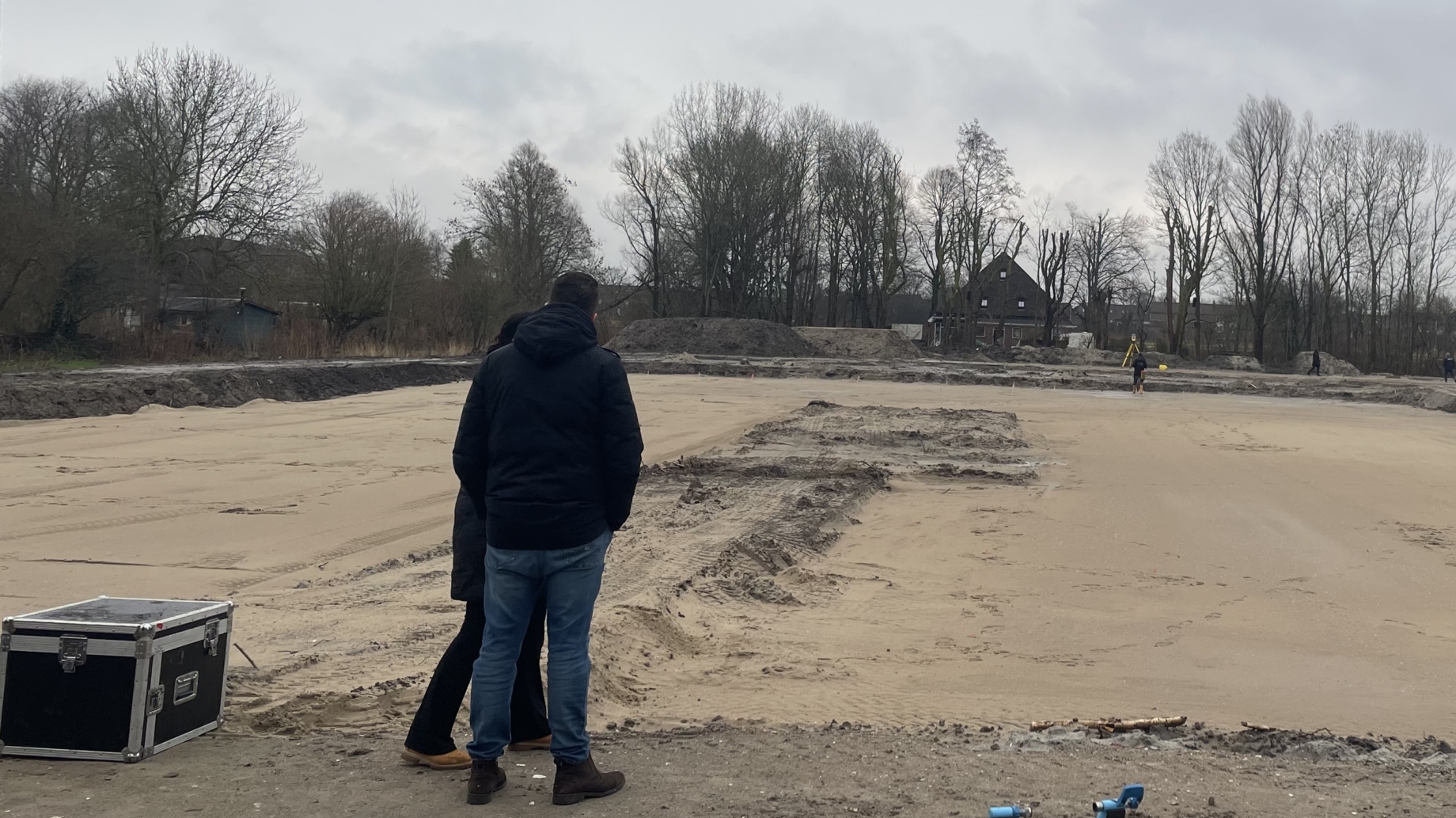 Mensen staan op een bouwplaats met een vlakke zandbodem en grijze lucht, omringd door bomen en gebouwen.